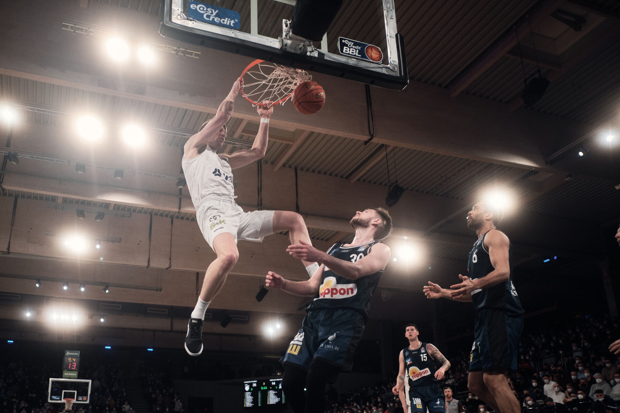 Basketballspieler in weißer Uniform macht Dunk während zwei Spieler in schwarzen Trikots versuchen zu verteidigen