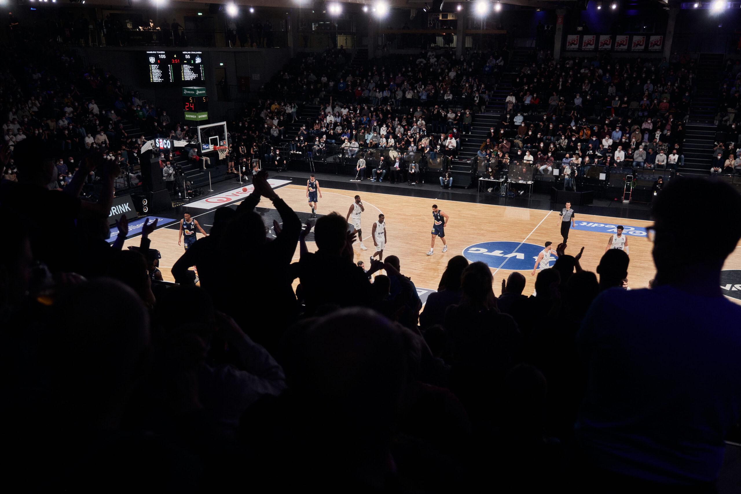 Basketballspiel in einer Halle mit Zuschauern, die im Vordergrund jubeln und klatschen