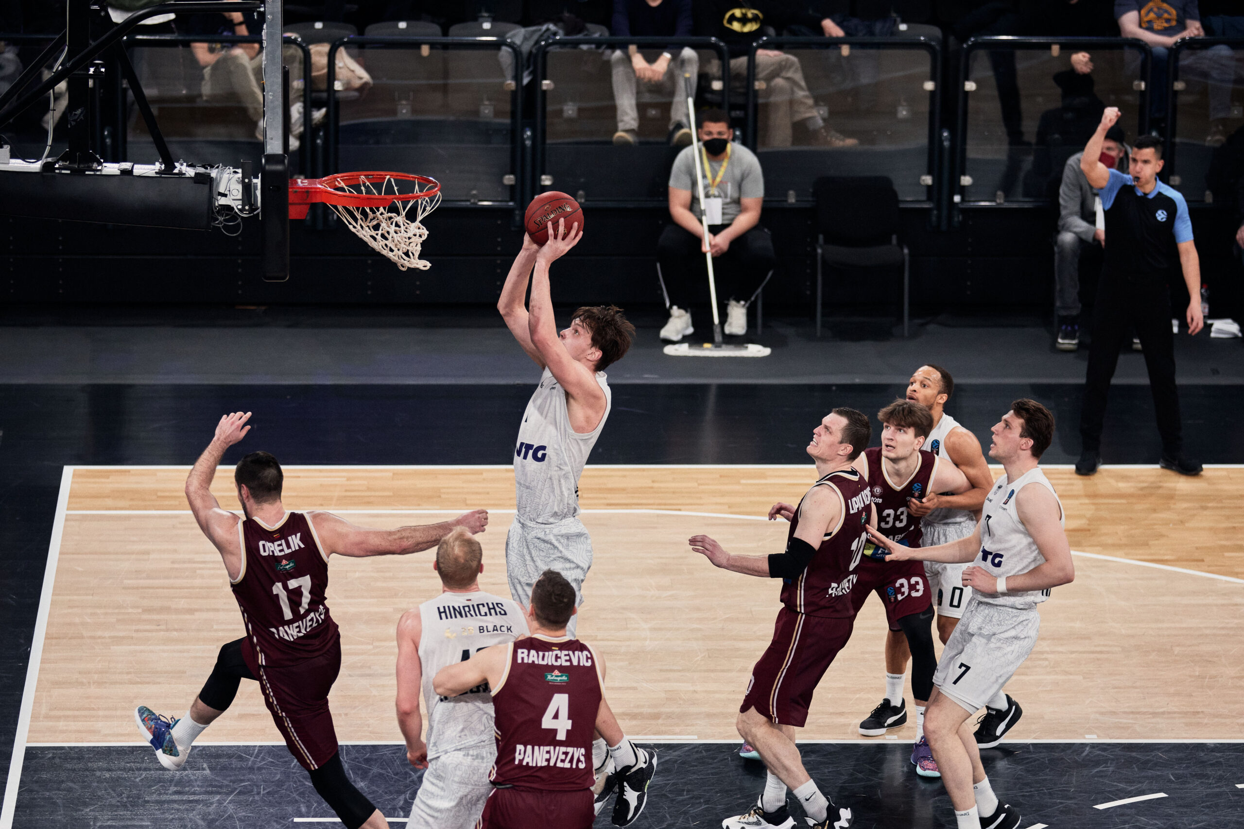 Basketballspieler in weißen Trikots beim Korbversuch, umgeben von Gegnern in dunkelroten Trikots auf dem Spielfeld