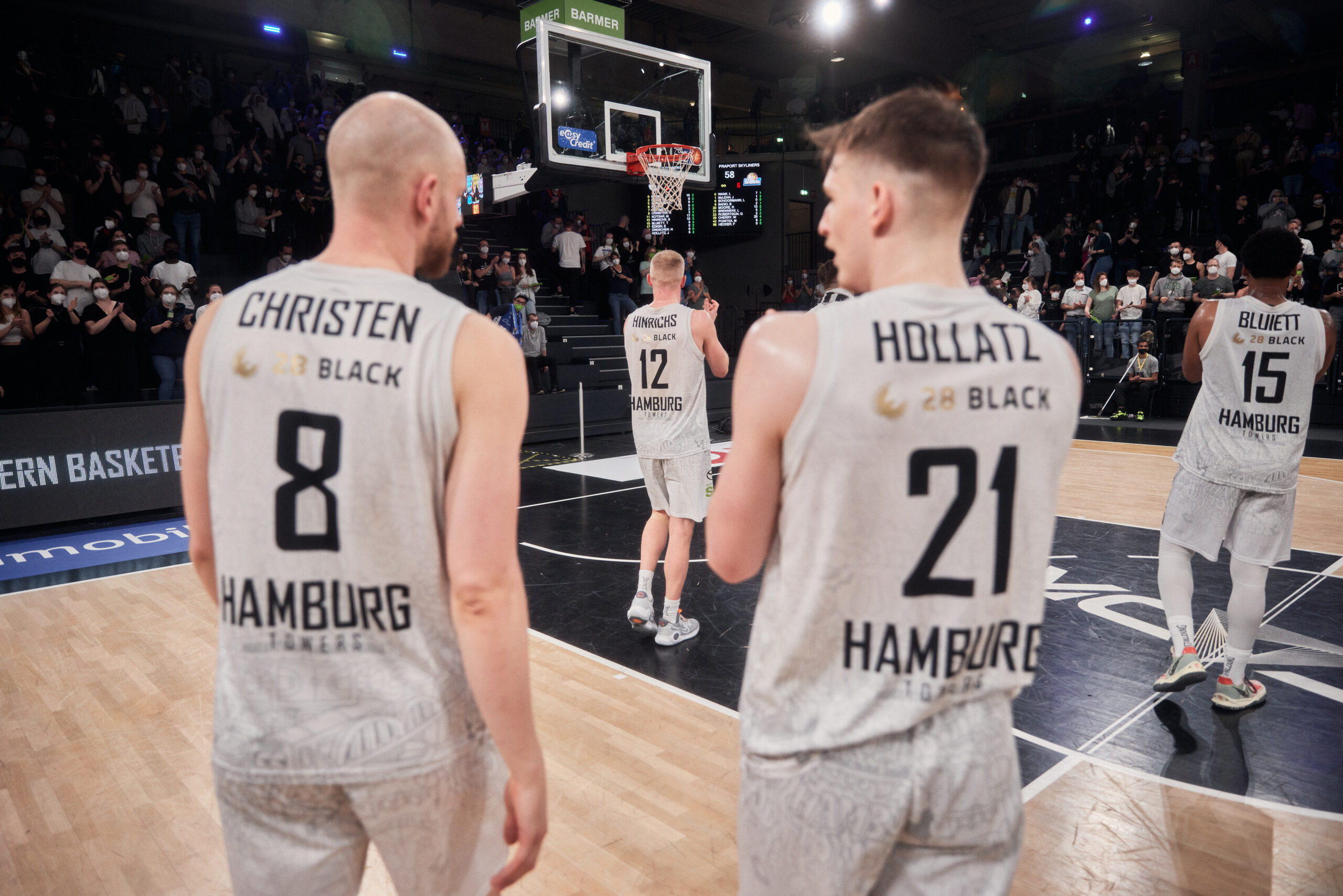 Vier Basketballspieler der Hamburg Towers in weißen Trikots mit den Nummern 8, 21, 12 und 15 auf dem Spielfeld vor einem Korb