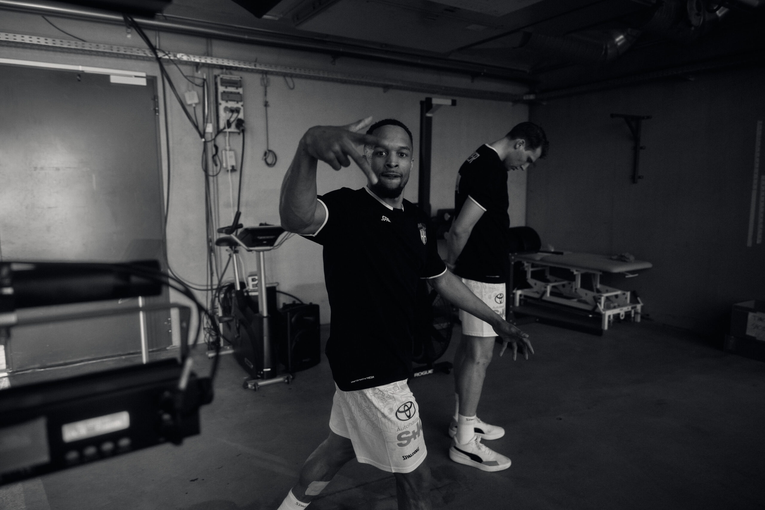 Zwei junge Männer in Sportkleidung in einem Innenraum, einer zeigt eine Handgeste zur Kamera, der andere schaut nach unten