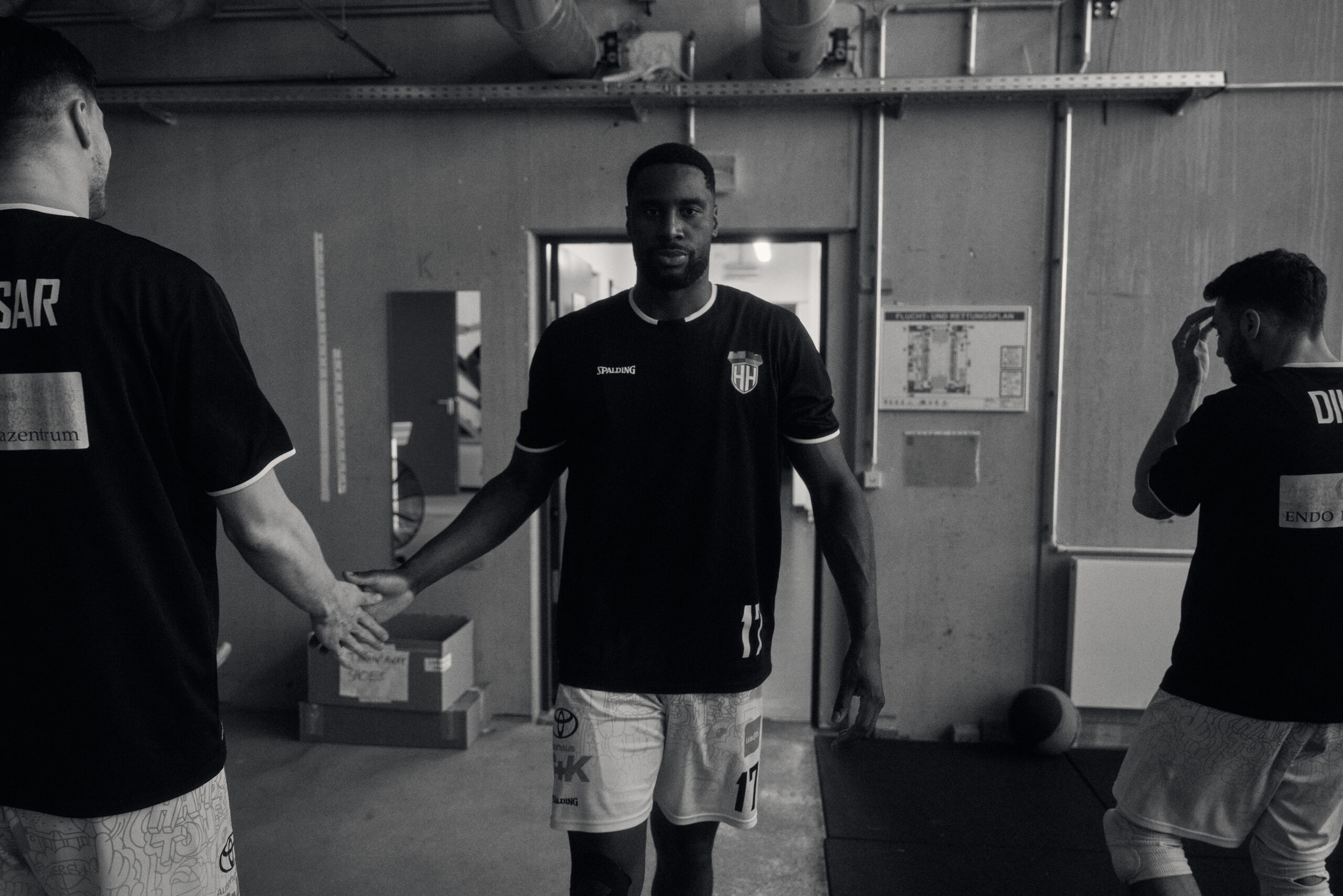 Drei Basketballspieler in Sportkleidung in einer Umkleide, einer in der Mitte gibt einem anderen die Hand.