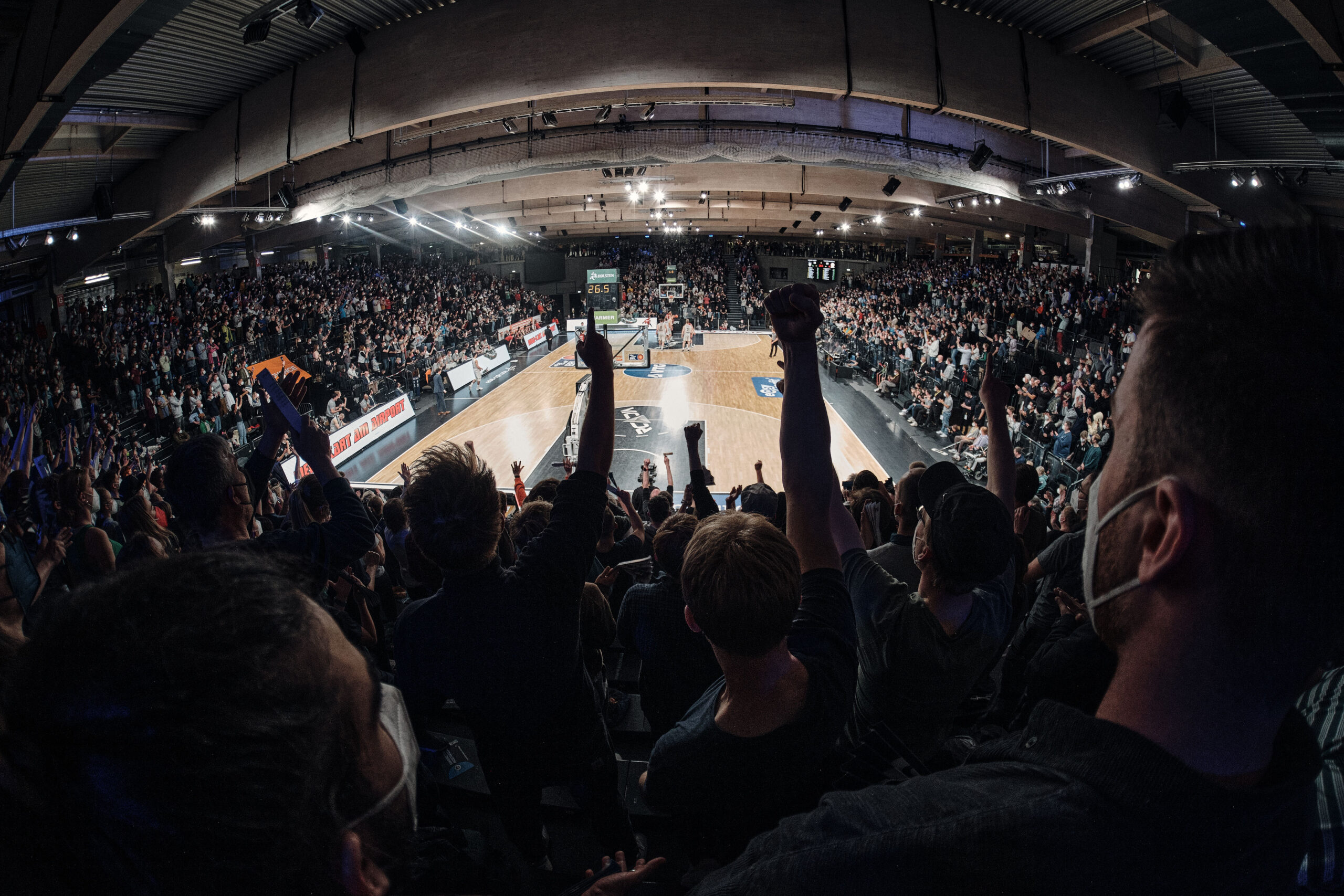 Voller Basketballarena mit jubelnden Zuschauern, die Arme heben, während ein Spiel auf dem Spielfeld läuft