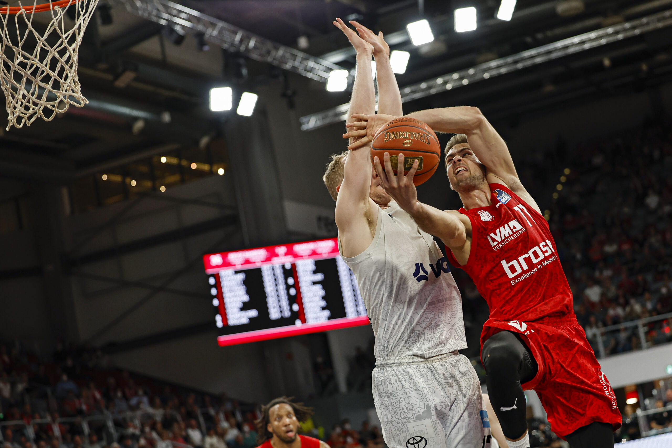 Zwei Basketballspieler kämpfen um den Ball nahe dem Korb, einer trägt rotes Trikot mit 'brose', der andere weißes Trikot, Spiel in Halle mit Zuschauertribüne