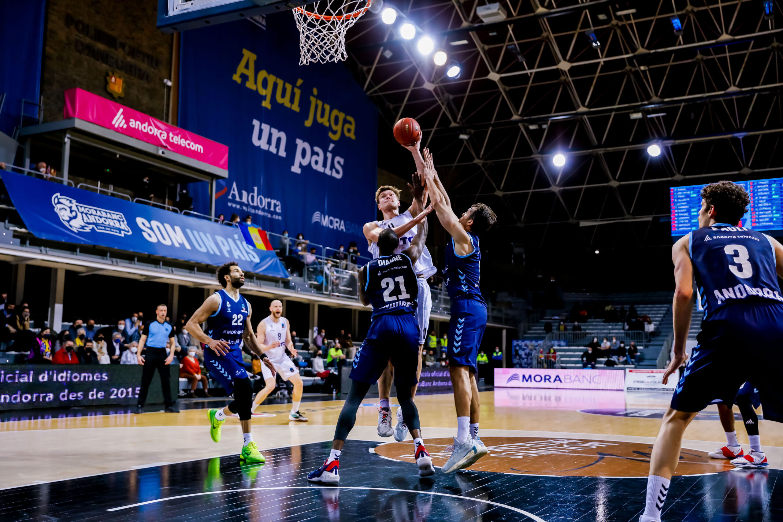 Basketballspieler in blauen Trikots verteidigen gegen einen Spieler in weißem Trikot, der zum Korb springt und den Ball hält