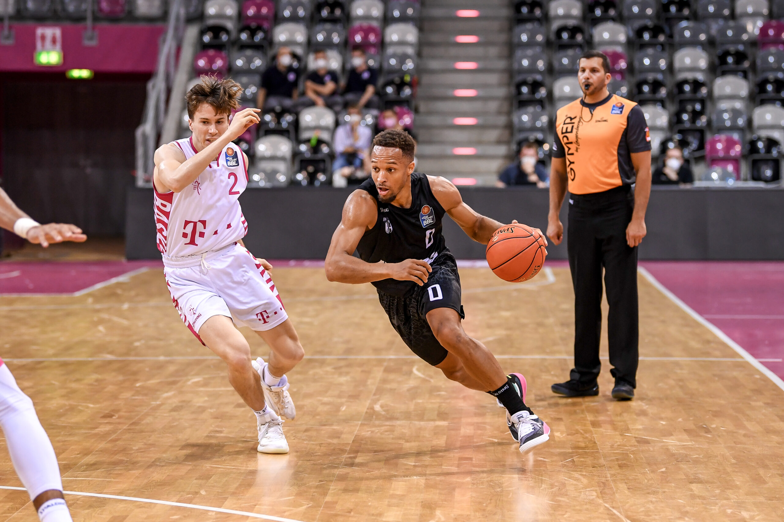 Basketballspieler in schwarzer Trikotfarbe dribbelt den Ball, während ein Spieler in weißem Trikot versucht, ihn zu verteidigen, Schiedsrichter beobachtet das Spiel