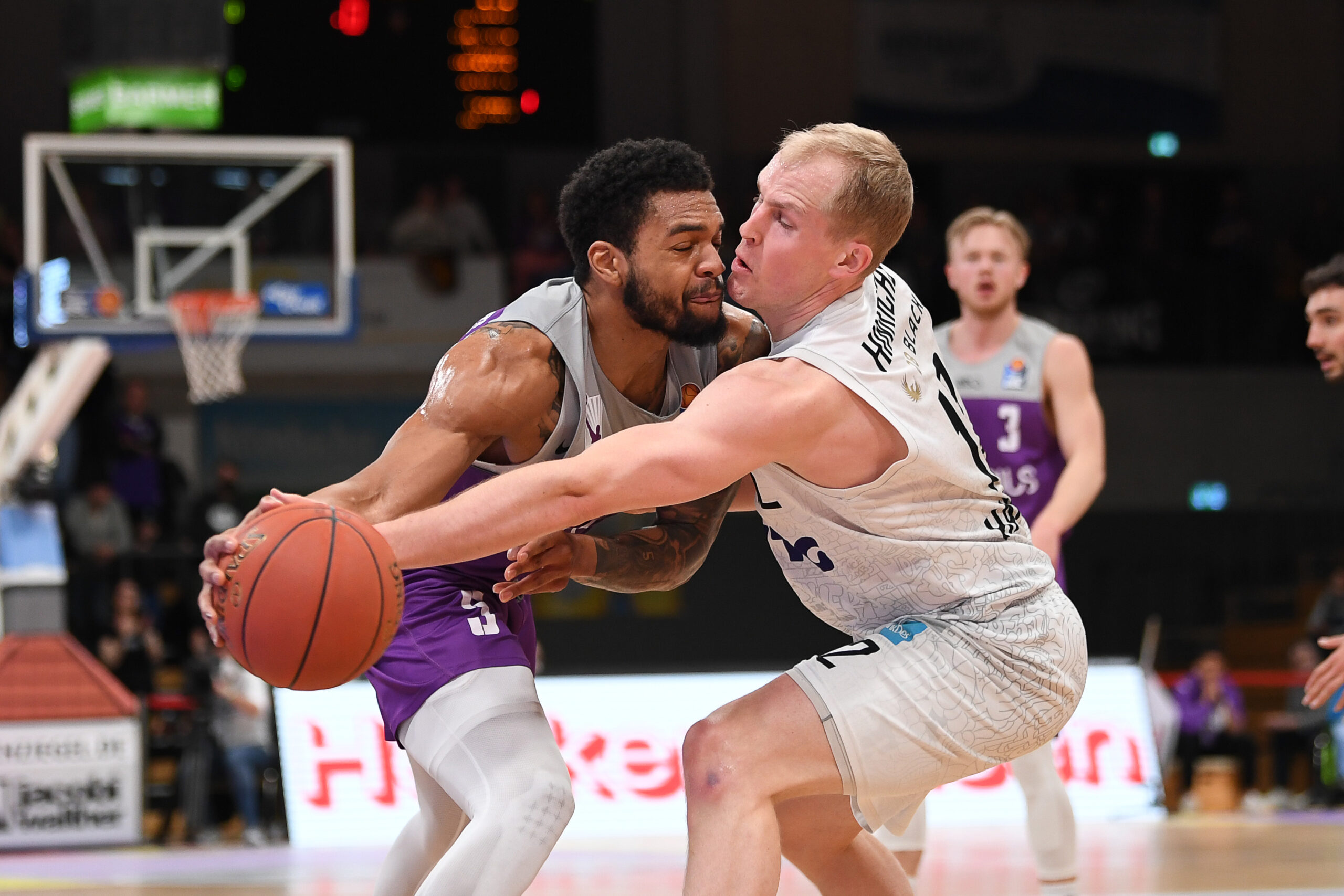 Zwei Basketballspieler kämpfen um den Ball, einer in weißem Trikot dribbelt, der andere in lila verteidigt intensiv.