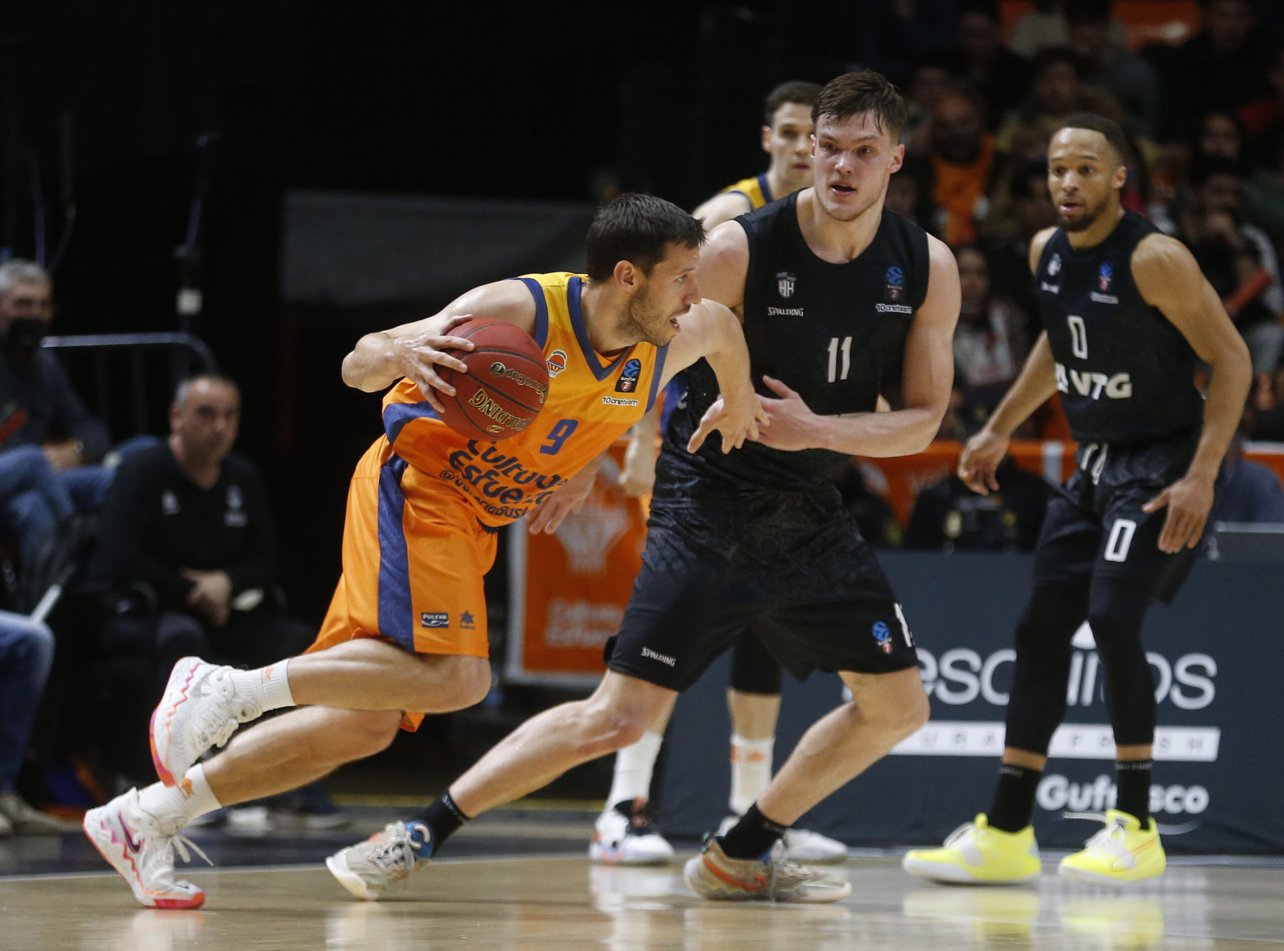 Basketballspieler in orangefarbenem Trikot dribbelt den Ball, während ein Spieler in schwarzem Trikot ihn verteidigt