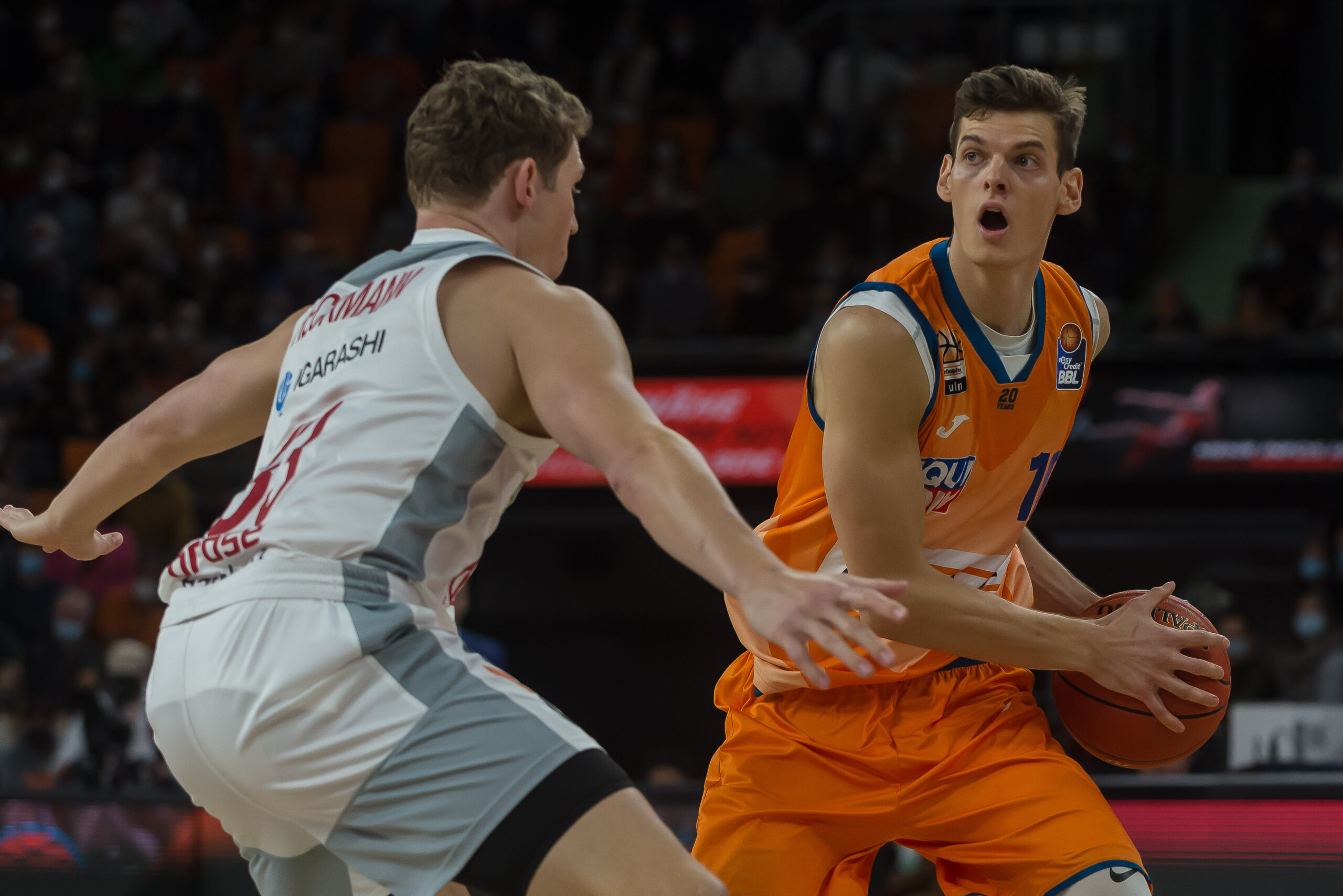 Basketballspieler in orange Trikots mit Ball, verteidigt von Spieler in weißem Trikot während eines Spiels