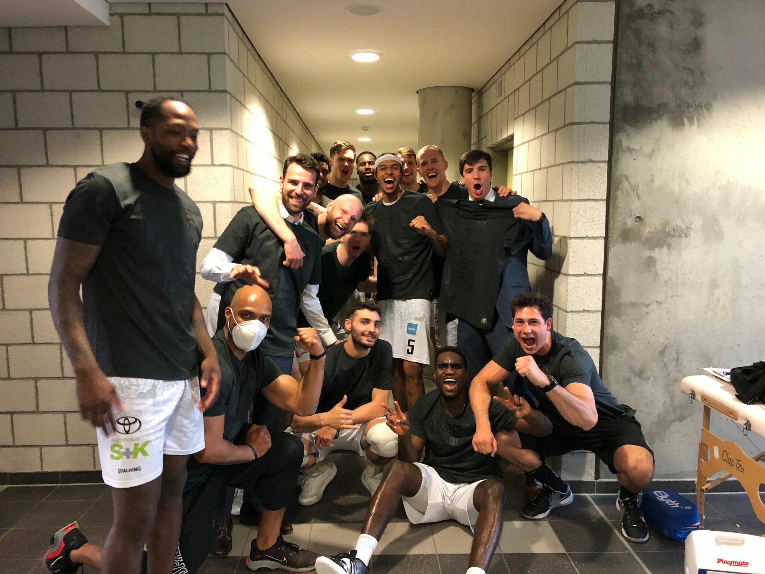 Gruppe von elf Männern in Sportkleidung, die in einem Flur zusammen lachen und posieren, einige machen Faustzeichen und zeigen Freude