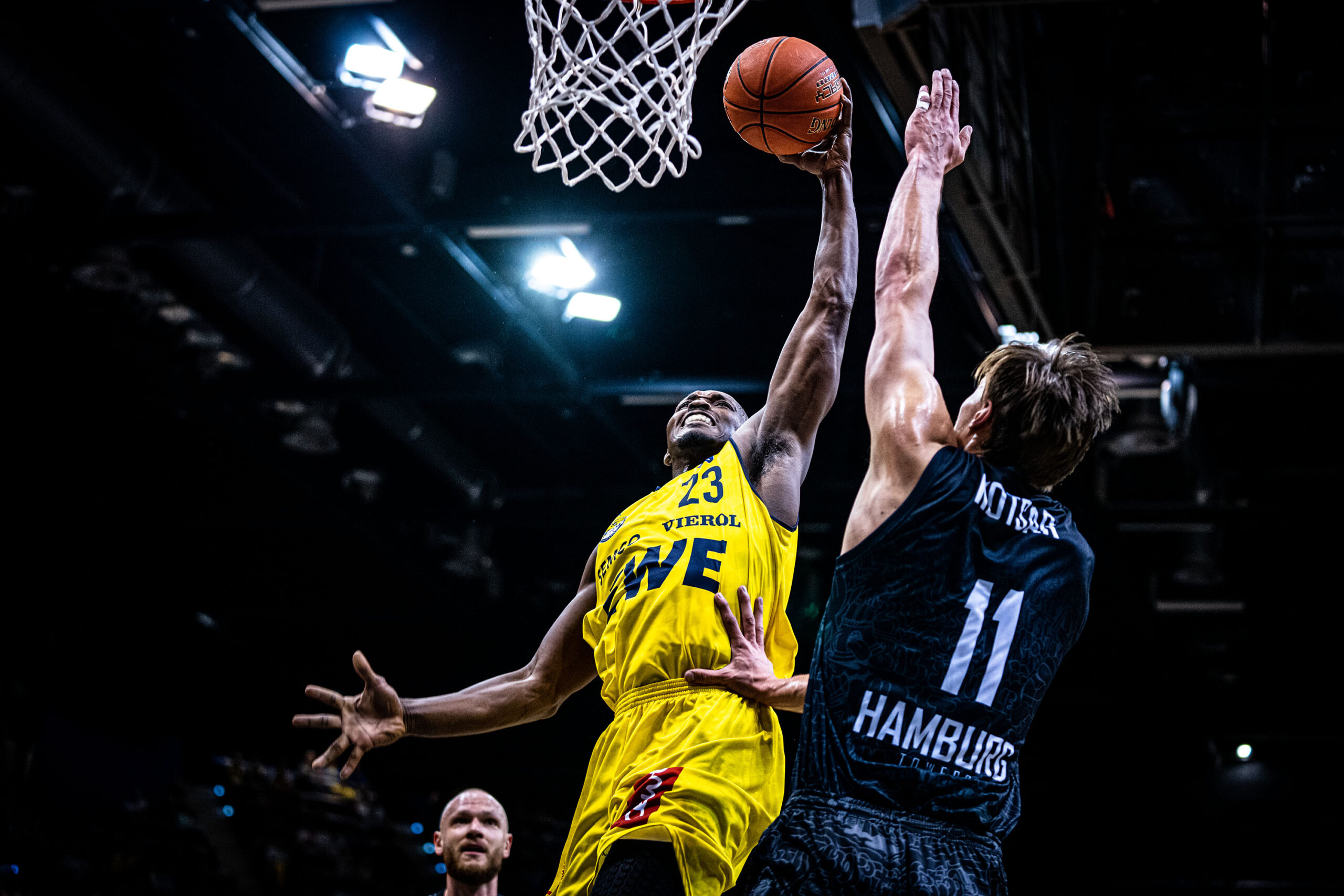 Basketballspieler in gelbem Trikot mit Ball beim Korbversuch, verteidigt von Spieler in schwarzem Trikot mit der Aufschrift 'Hamburg'