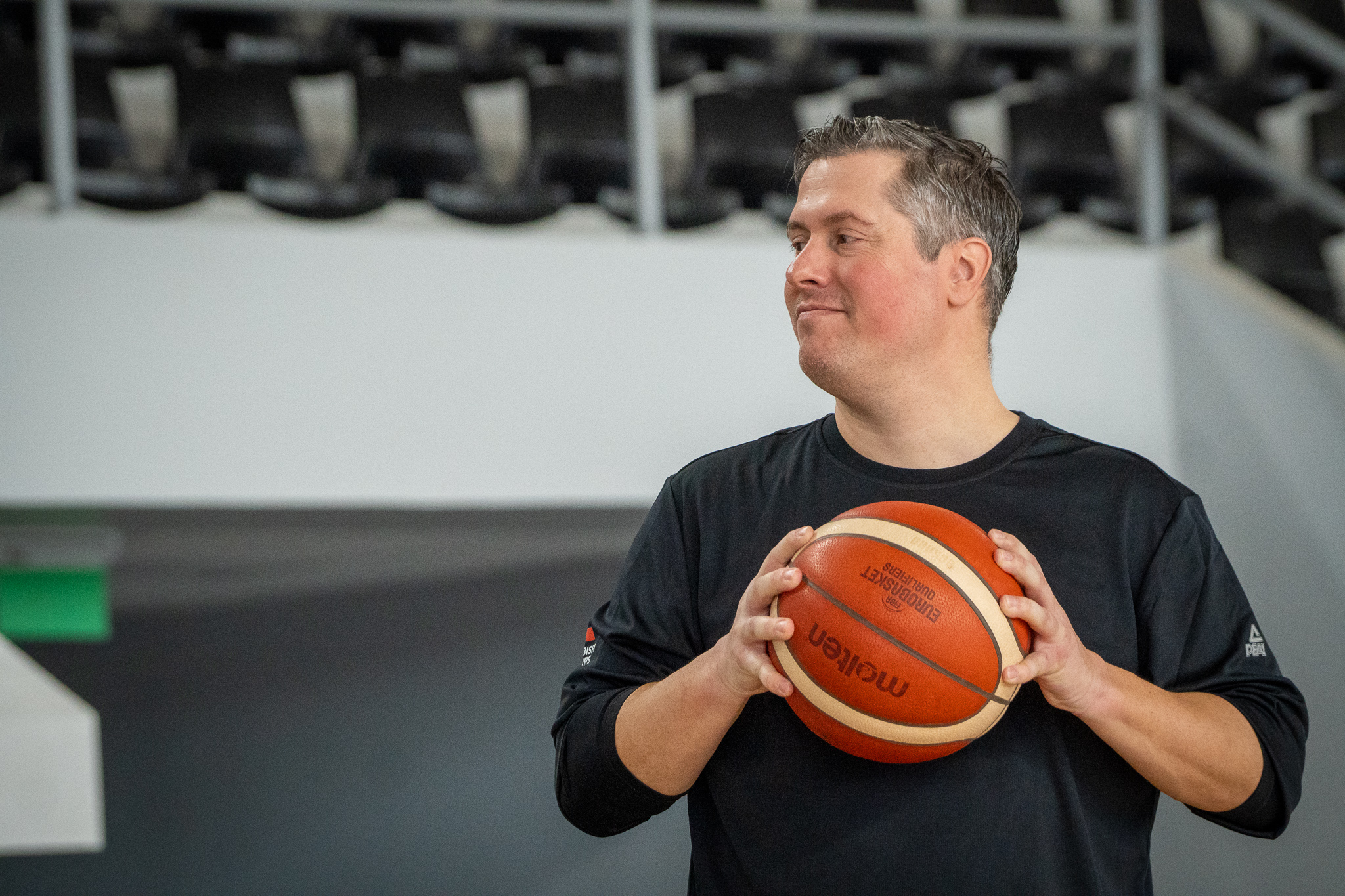 Mann in schwarzem Shirt hält einen Basketball und schaut nach rechts in einer Sporthalle