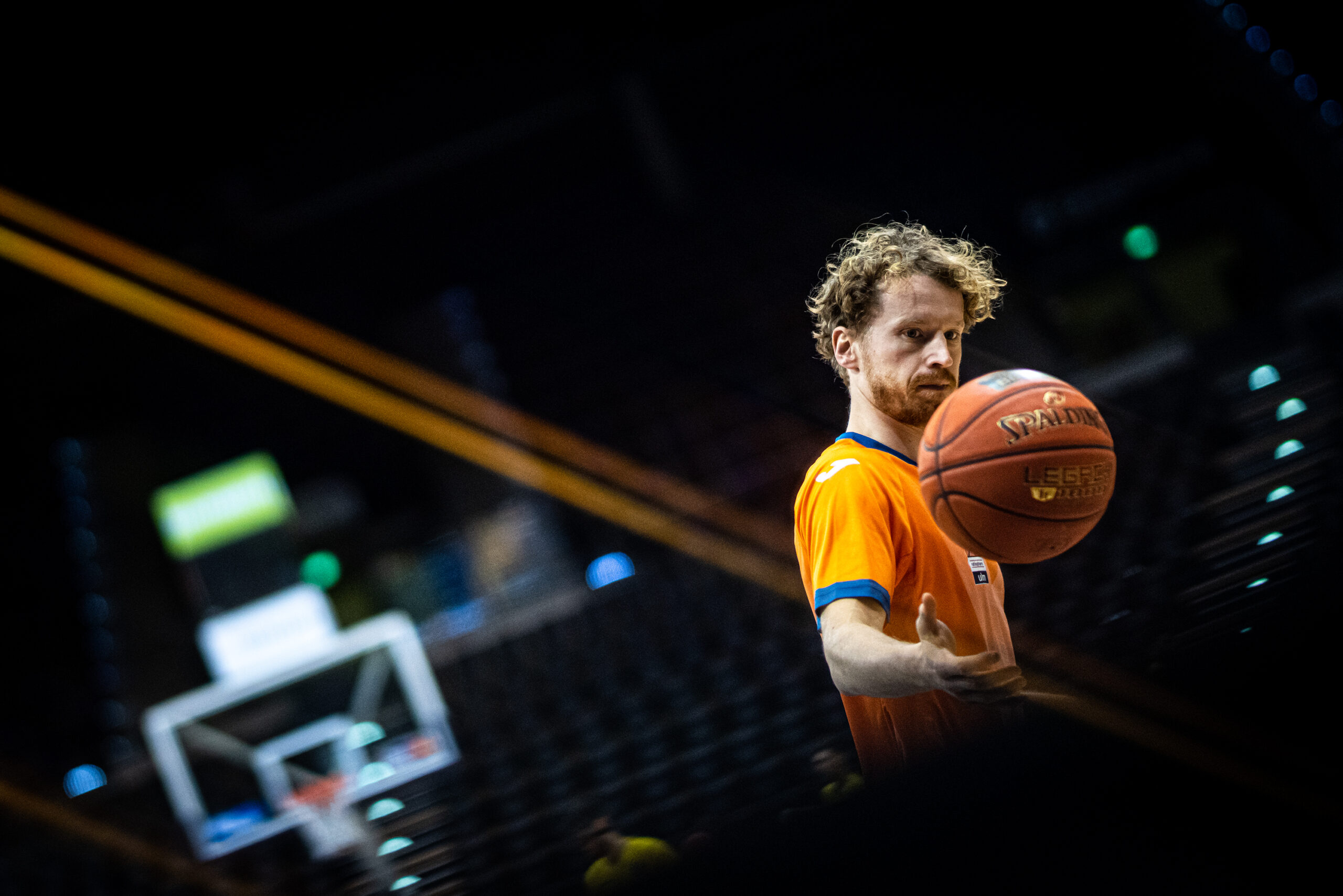Mann in orangefarbenem Trikot wirft Basketball in einer Halle mit Basketballkorb im Hintergrund