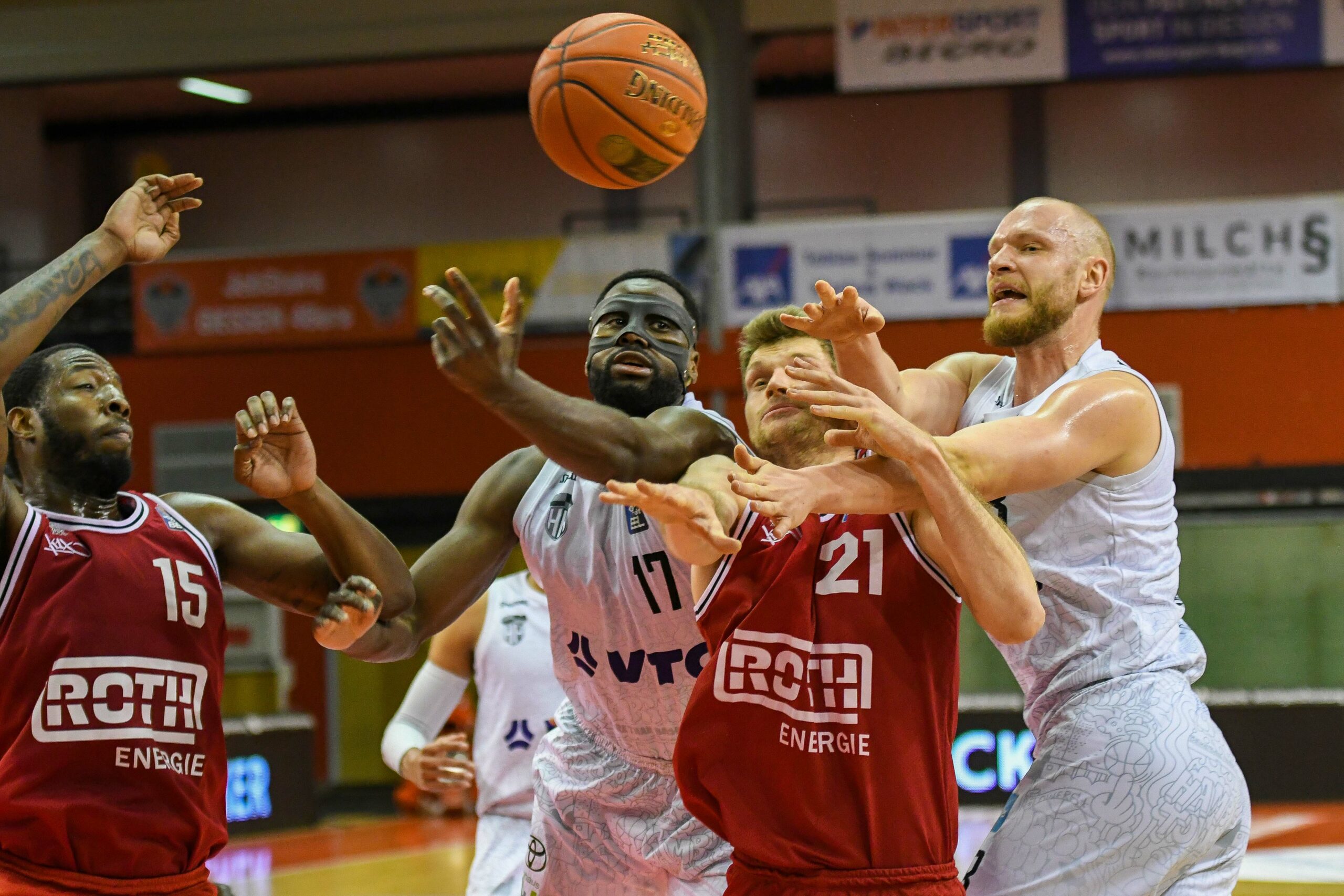 Basketballspieler kämpfen um den Ball in einem Spiel, zwei Spieler in weißen Trikots und zwei in roten Trikots mit der Aufschrift 'ROTH ENERGIE'