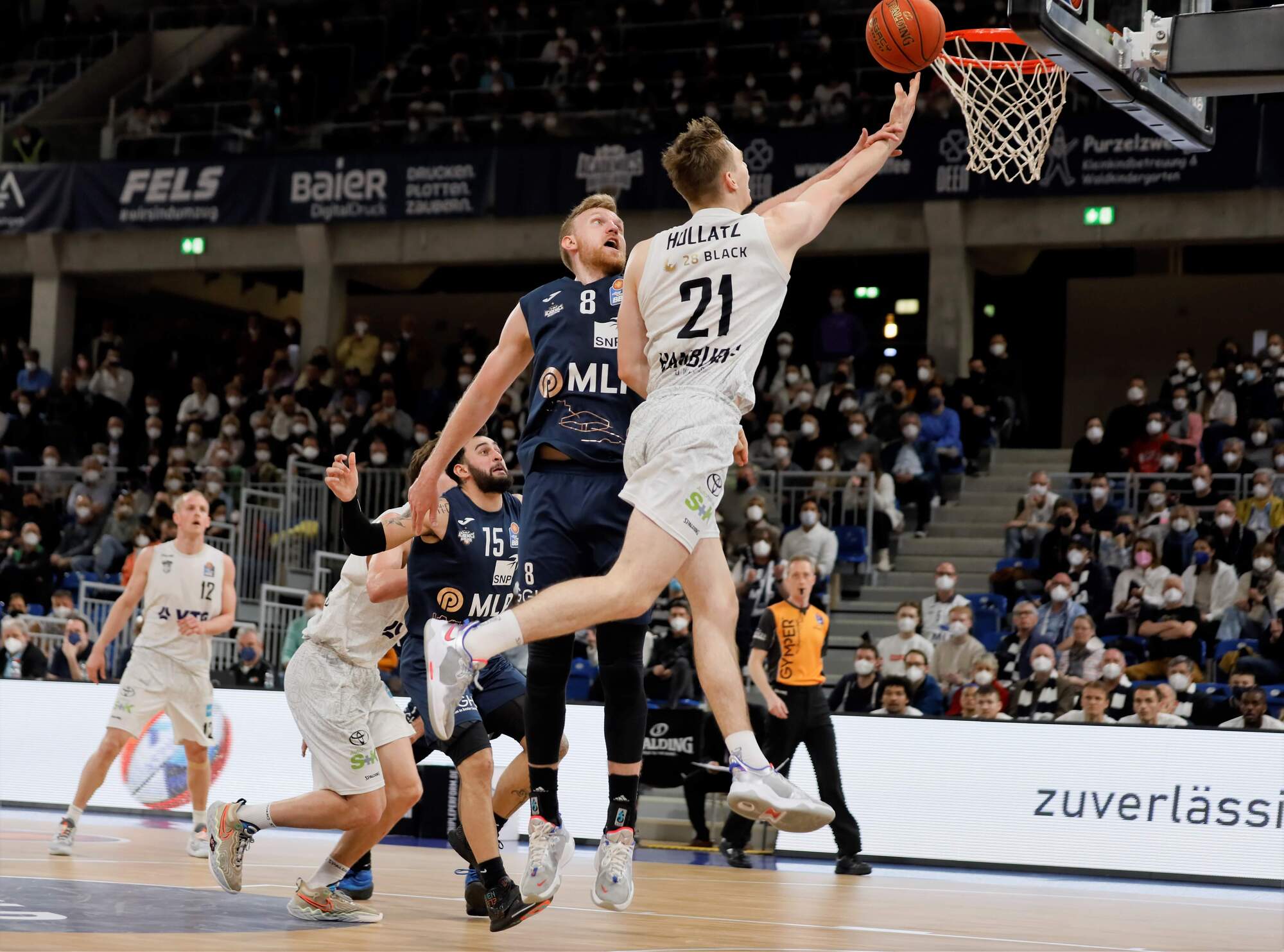Basketballspieler mit Trikotnummer 21 springt zum Korb für einen Layup, während ein Verteidiger versucht zu blocken, Zuschauer im Hintergrund