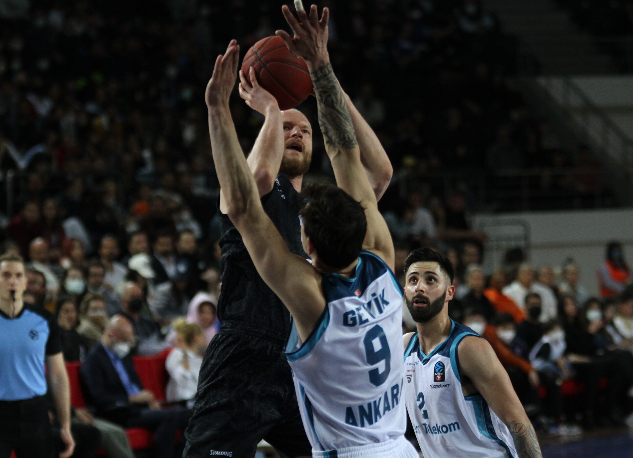 Basketballspieler in schwarzem Trikot wirft Ball, während zwei Spieler in weißen Trikots mit der Aufschrift 'Ankara' und Nummern 9 und 3 verteidigen