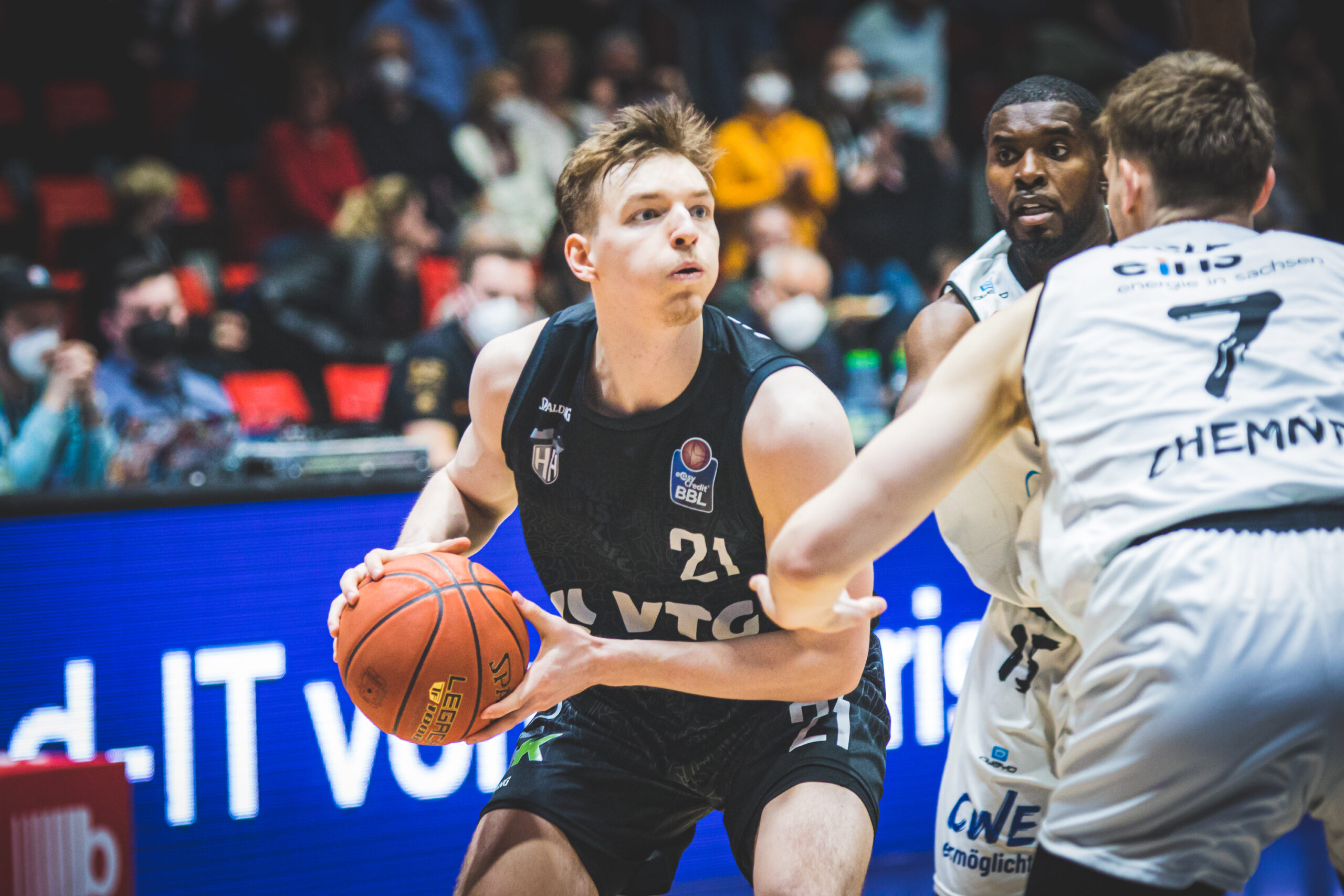 Basketballspieler in schwarzem Trikot mit Ball, umgeben von zwei verteidigenden Spielern in weißen Trikots während eines Spiels