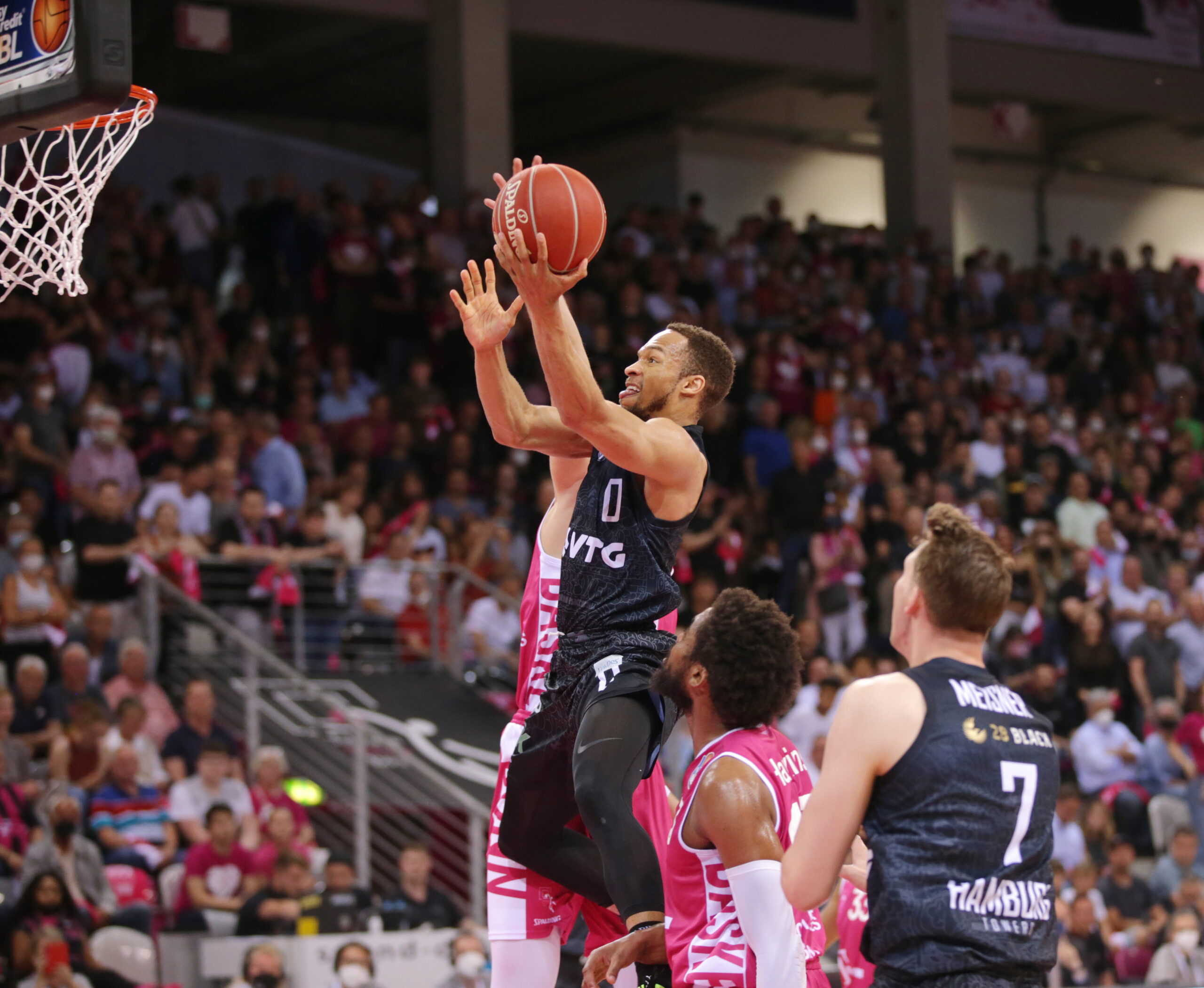 Basketballspieler in dunkler Uniform springt zum Korb, um einen Korb zu erzielen, umgeben von Gegnern in pinken Trikots, Zuschauer im Hintergrund