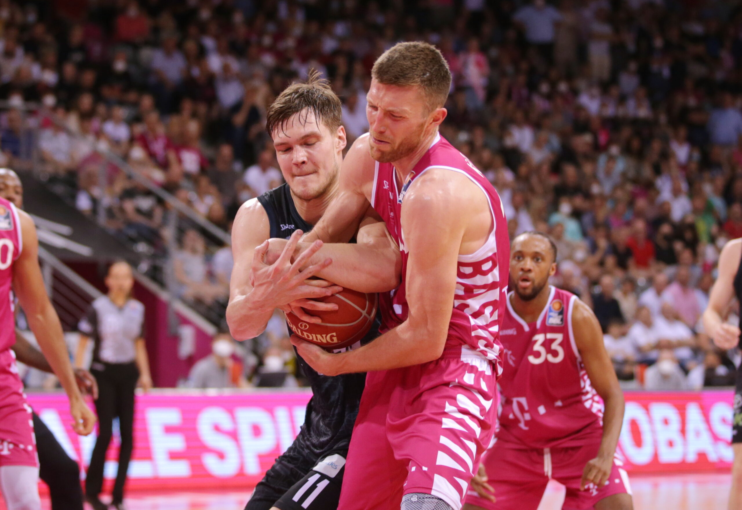 Zwei Basketballspieler kämpfen um den Ball, einer trägt schwarzen Trikot, der andere pinkfarbenes Trikot, Zuschauer im Hintergrund