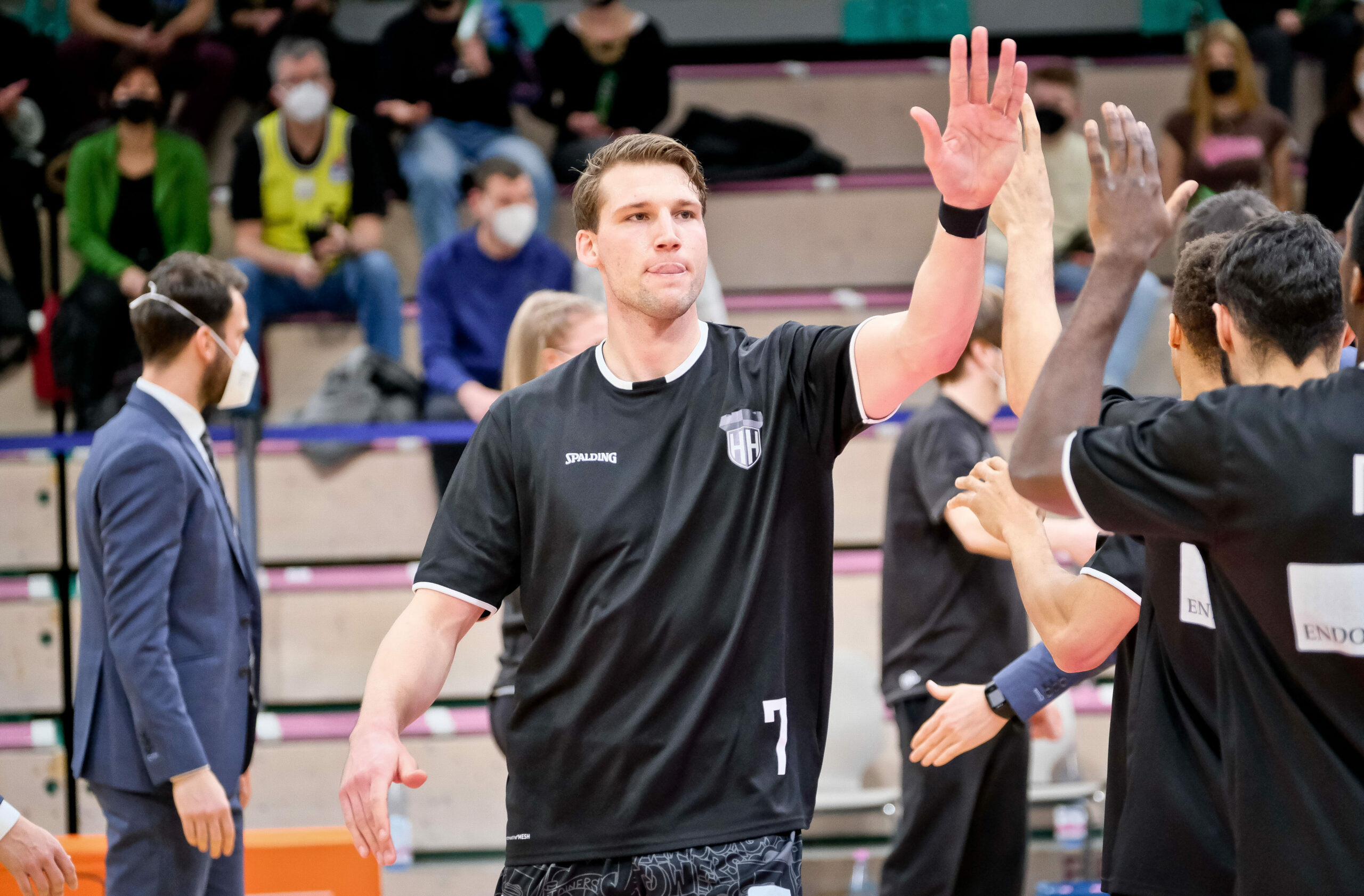 Basketballspieler in schwarzem Trikot mit der Nummer 7 gibt einem Teamkollegen einen High-Five während eines Spiels mit Zuschauern im Hintergrund