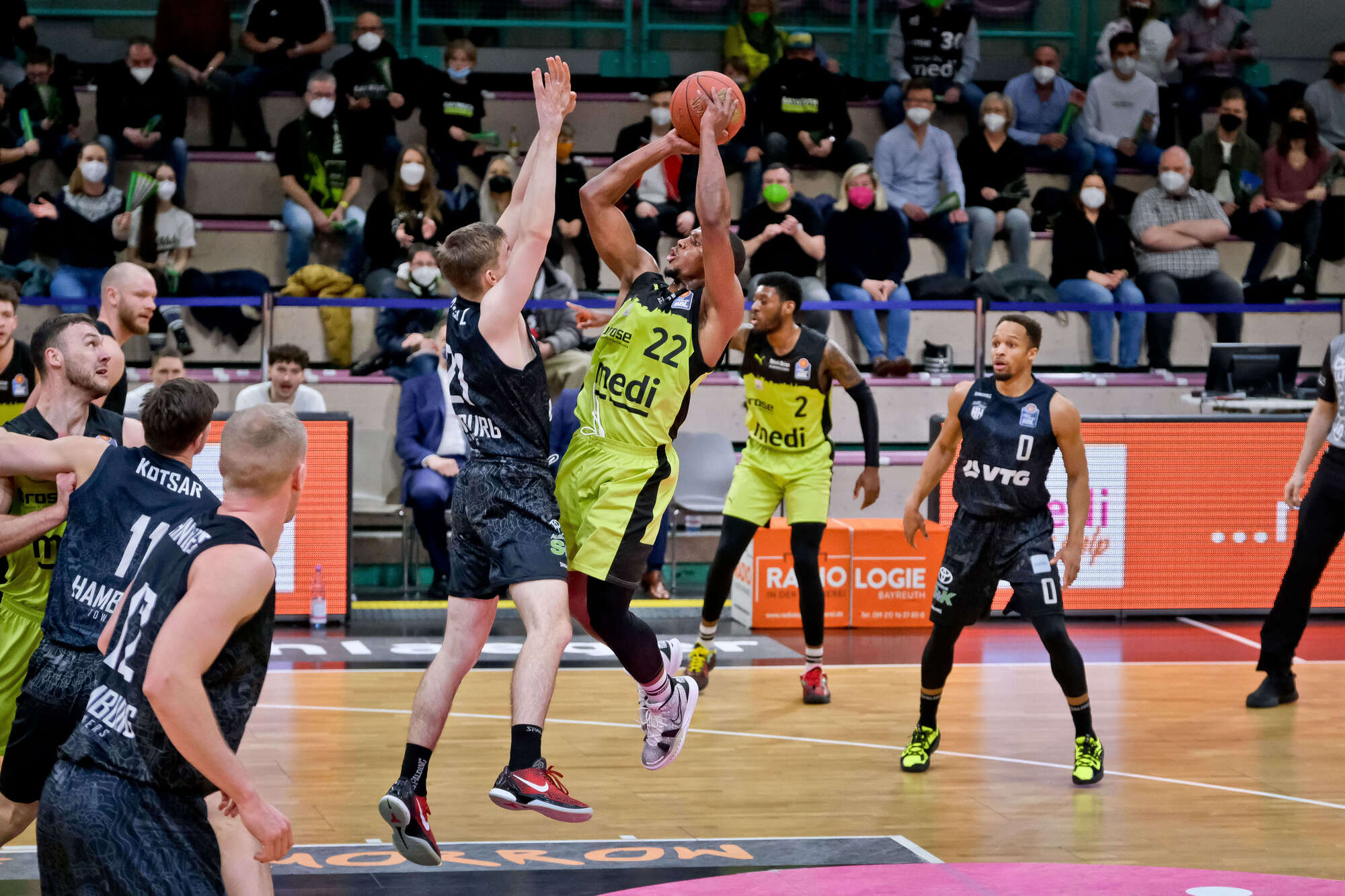 Basketballspieler in grünen Trikots beim Wurfversuch, verteidigt von Spielern in schwarzen Trikots, Zuschauer im Hintergrund