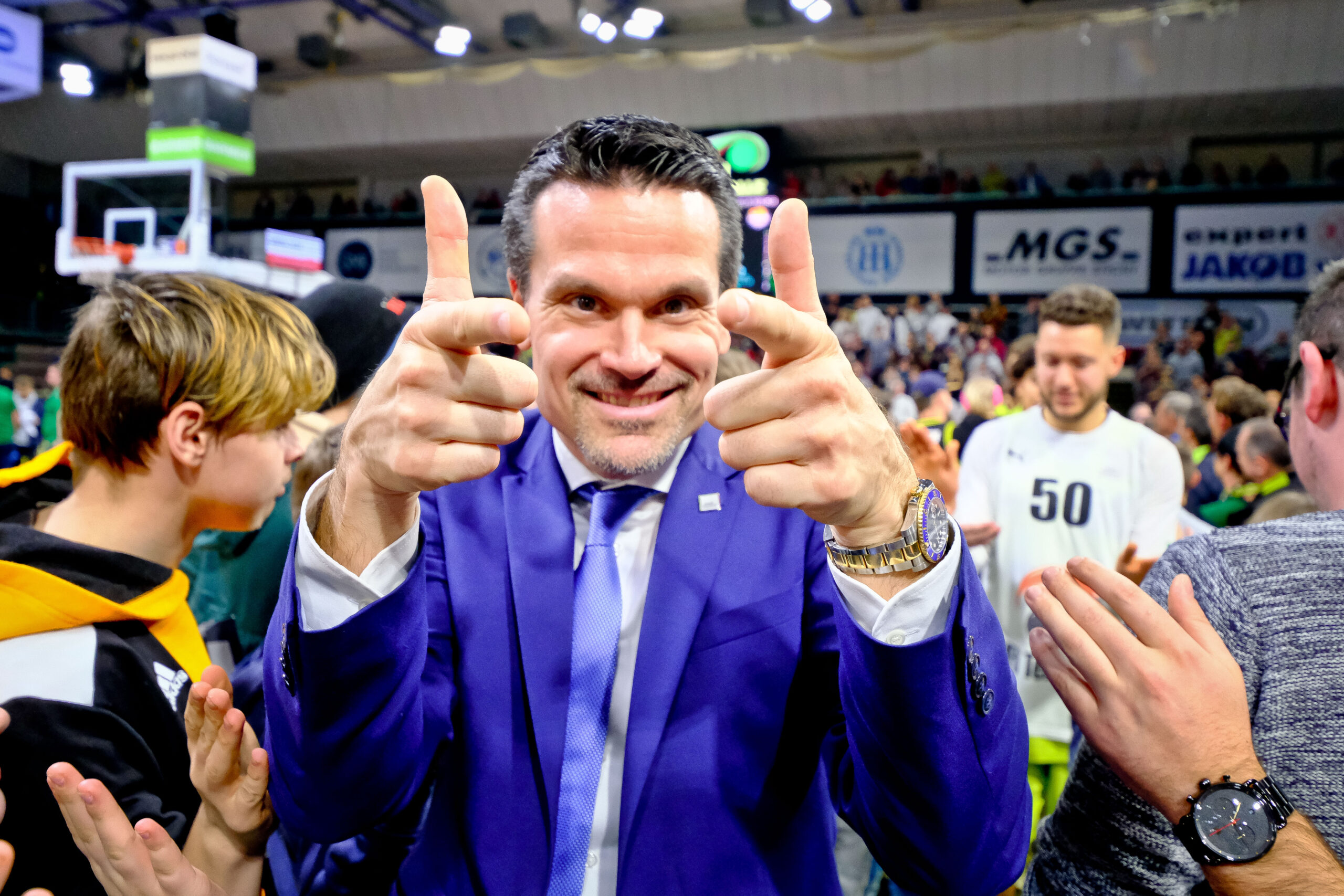 Mann im blauen Anzug zeigt mit beiden Händen Fingerpistolen und lächelt in einer Sporthalle mit jubelndem Publikum