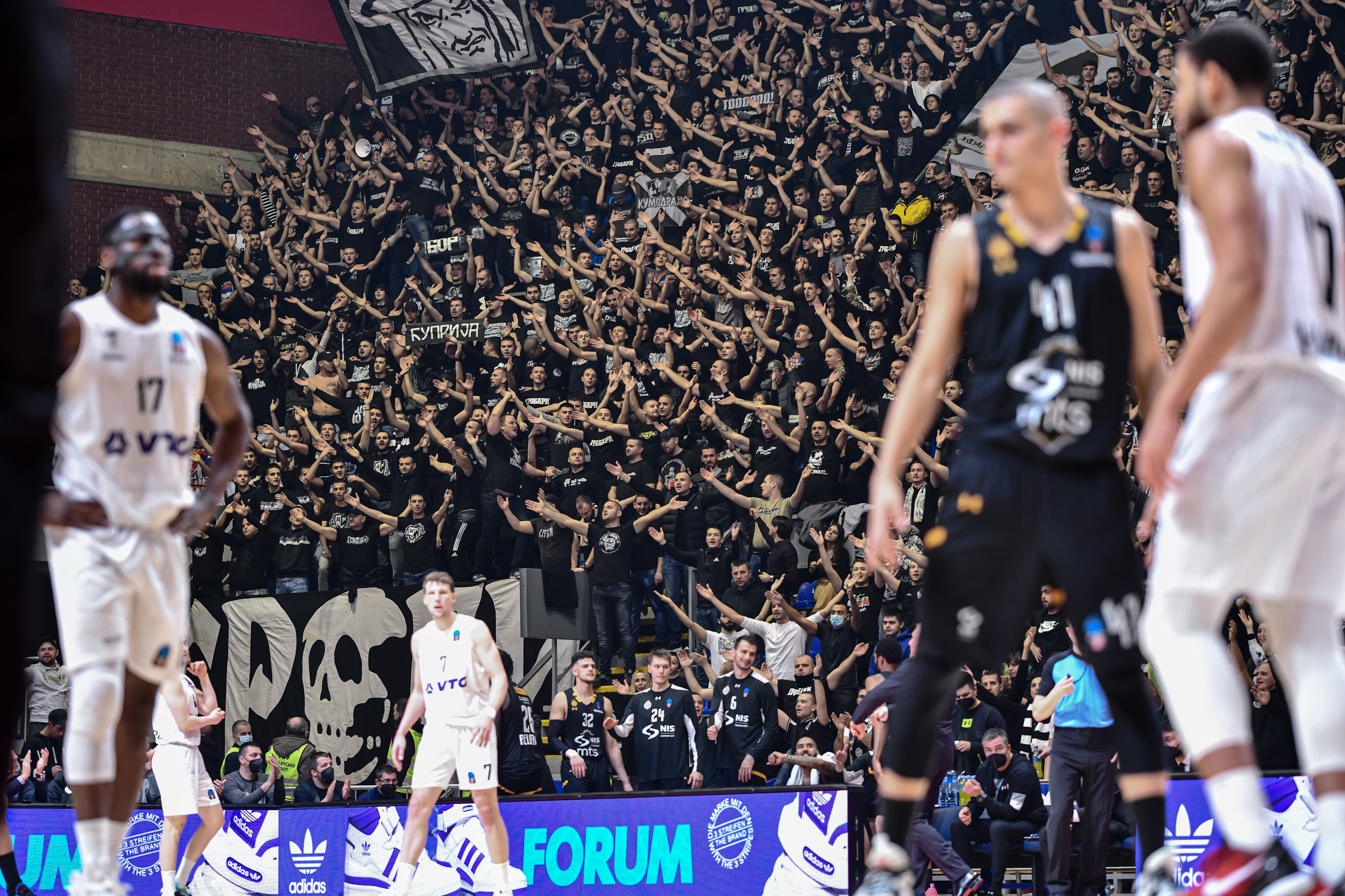 Basketballspiel mit Spielern in weißen und schwarzen Trikots vor einer Tribüne voller jubelnder Fans in schwarzer Kleidung