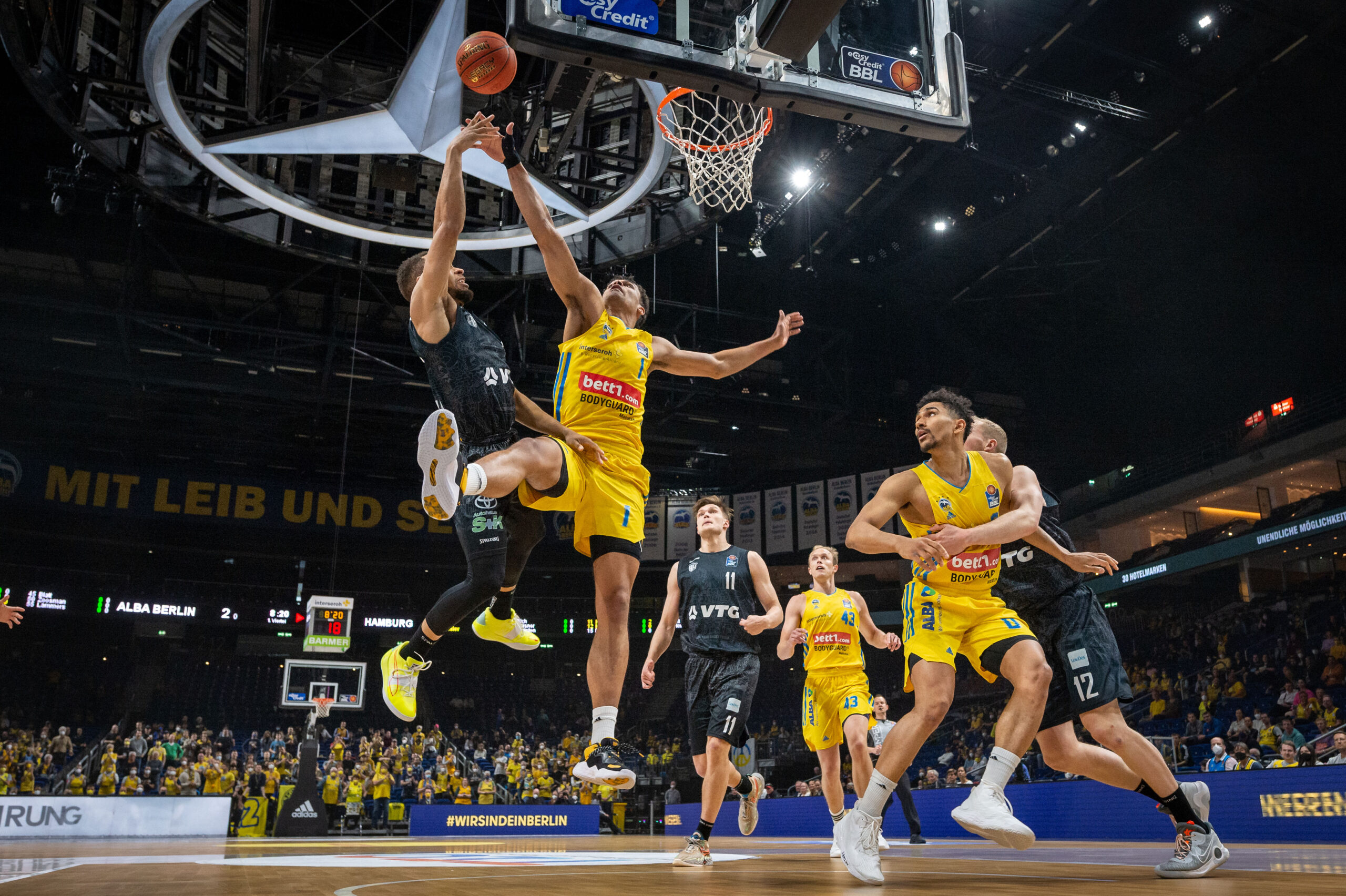 Basketballspieler in gelben und schwarzen Trikots kämpfen um den Ball unter dem Korb in einer vollen Halle während eines Spiels