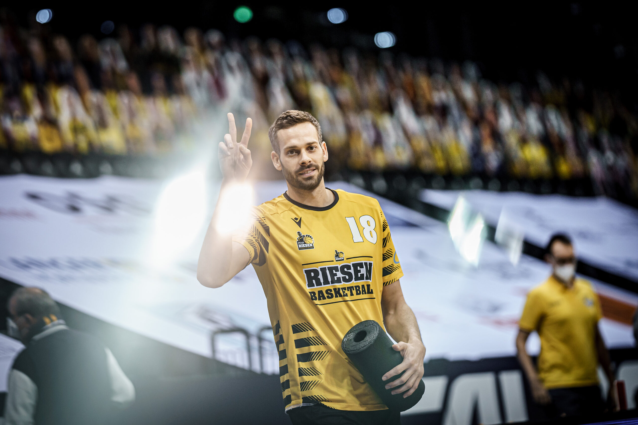 Basketballspieler in gelbem Trikot mit der Nummer 18 zeigt das Victory-Zeichen und hält eine Schaumstoffrolle in der Hand