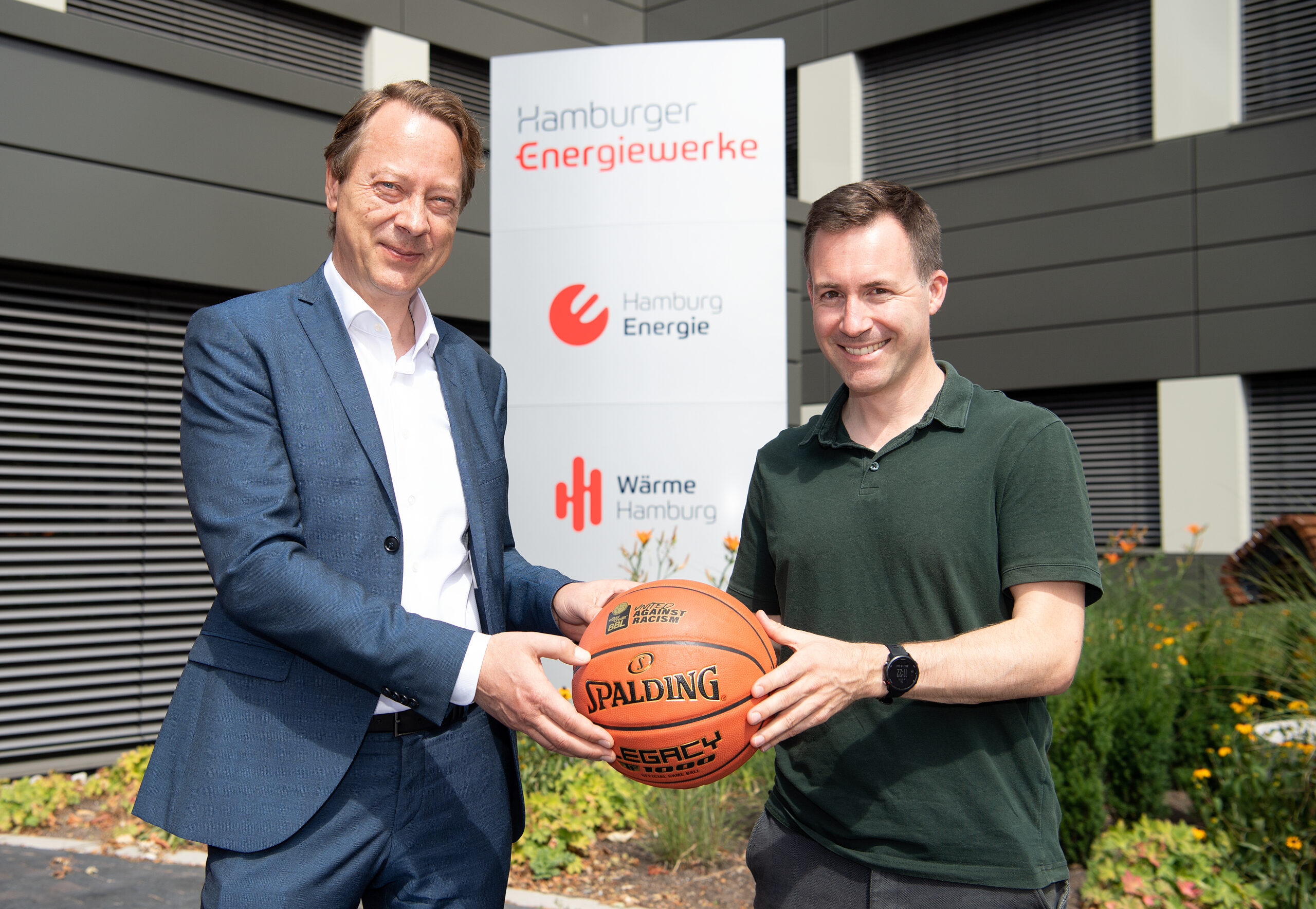 Zwei Männer vor einem Schild der Hamburger Energiewerke, die gemeinsam einen Basketball halten und lächeln.