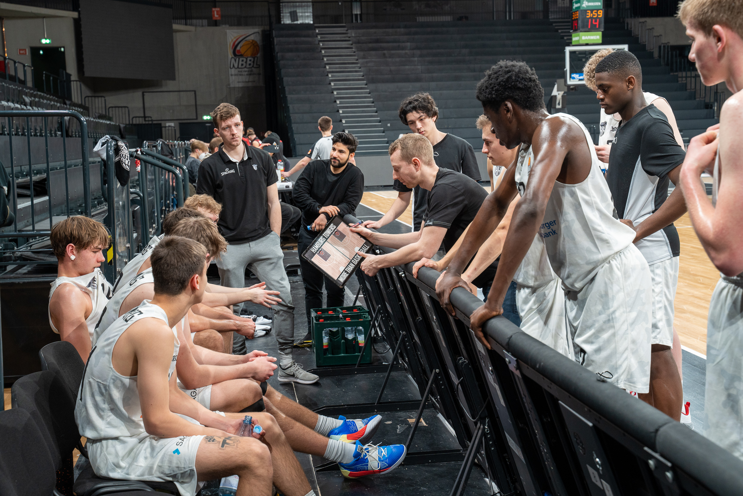 Basketballteam in weißen Trikots sitzt und steht um Trainer, der Taktik auf Tafel zeigt, in einer Sporthalle