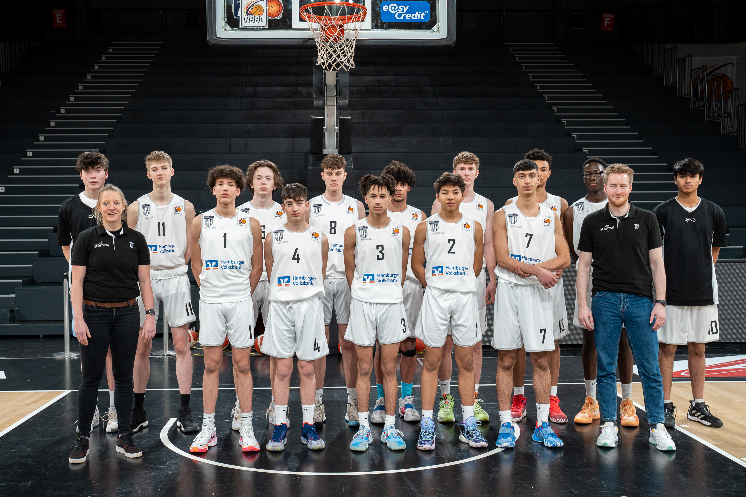 Jugend-Basketballmannschaft in weißen Trikots steht unter einem Basketballkorb in einer Halle, Trainer und Spieler in einer Reihe