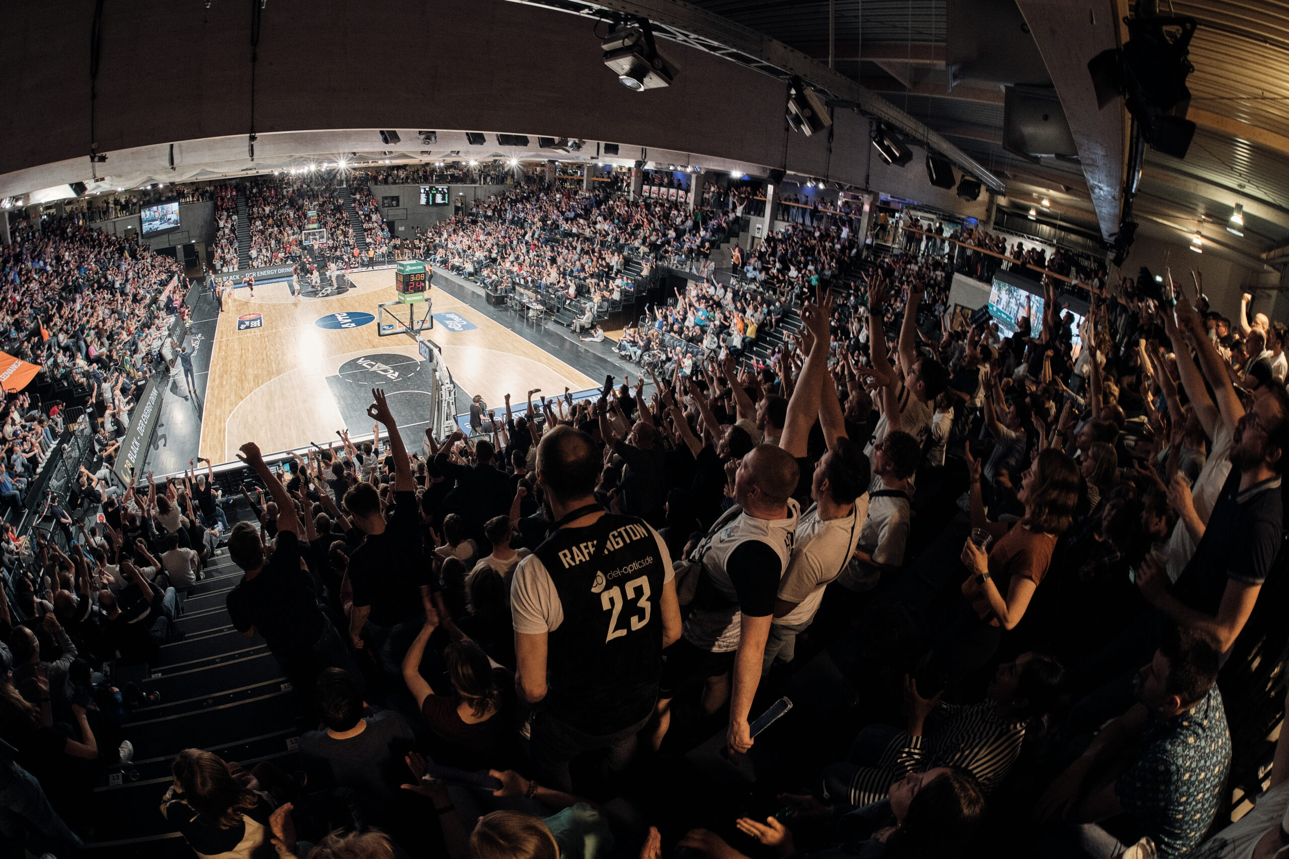 Voller Basketballarena mit jubelnden Zuschauern, die Arme heben, während ein Spiel auf dem Spielfeld läuft