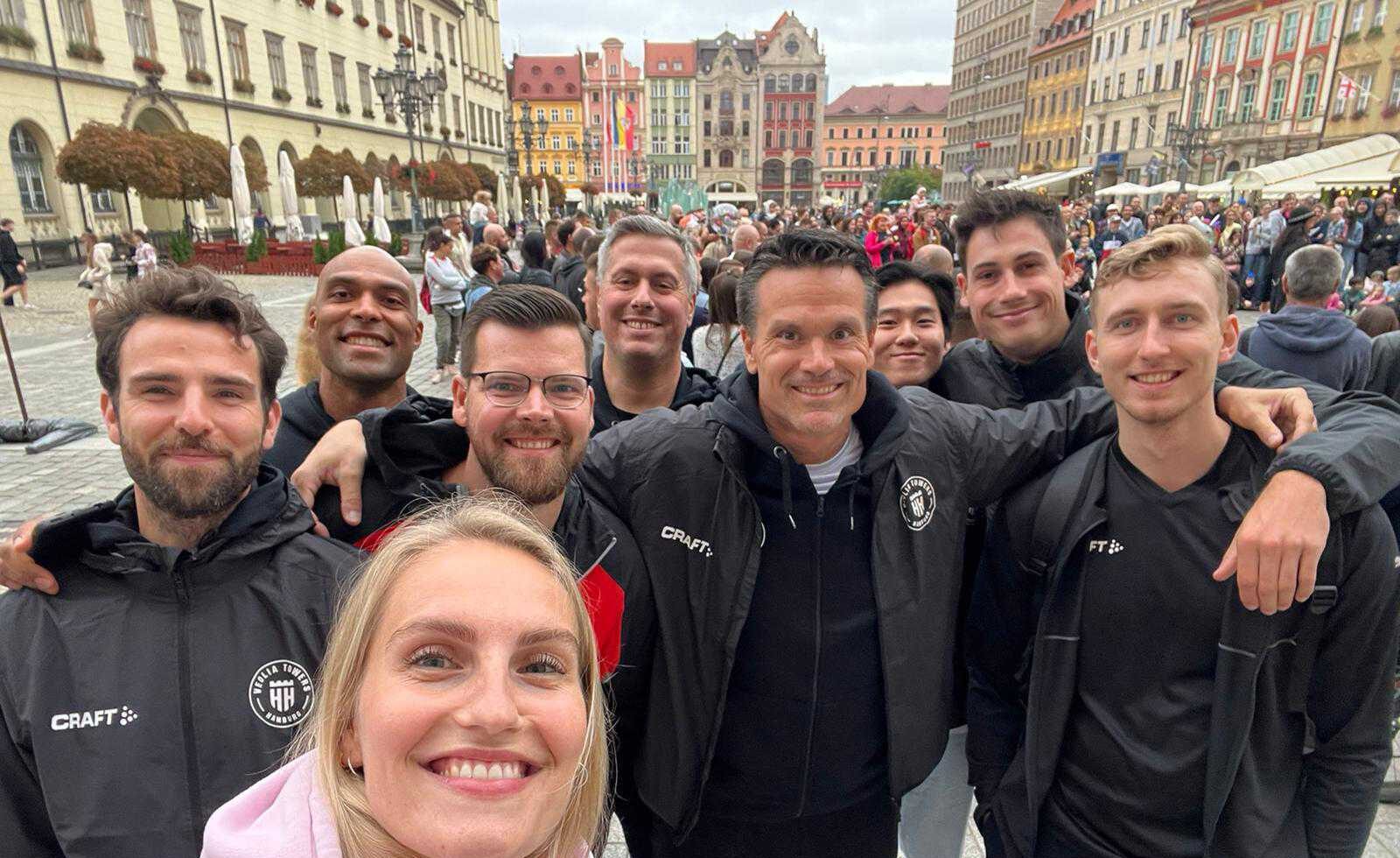Gruppe von neun Personen, die in einer belebten Stadt mit historischen Gebäuden im Hintergrund lächeln und eine Selfieaufnahme machen.