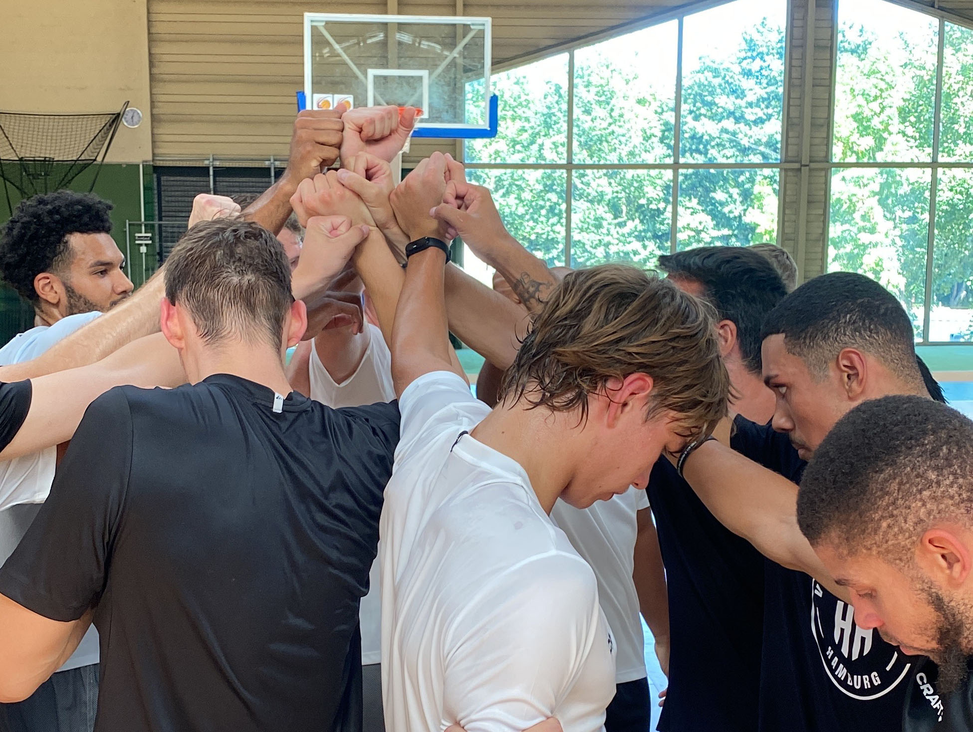Basketballteam in Sportkleidung bildet Teamkreis mit erhobenen Händen in einer Halle mit Basketballkorb
