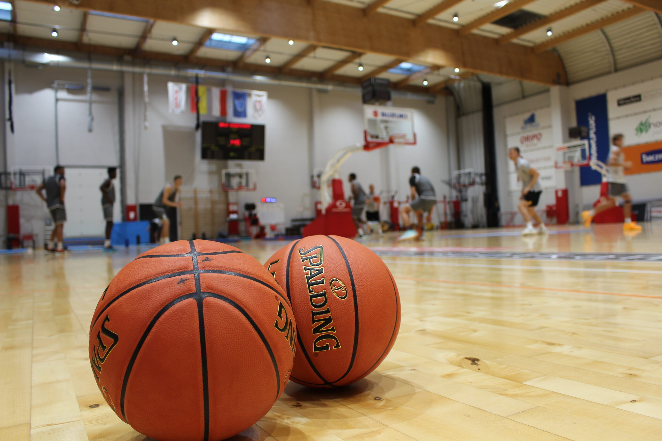Zwei orangefarbene Basketballs auf dem Holzboden einer Halle mit Spielern und Körben im Hintergrund