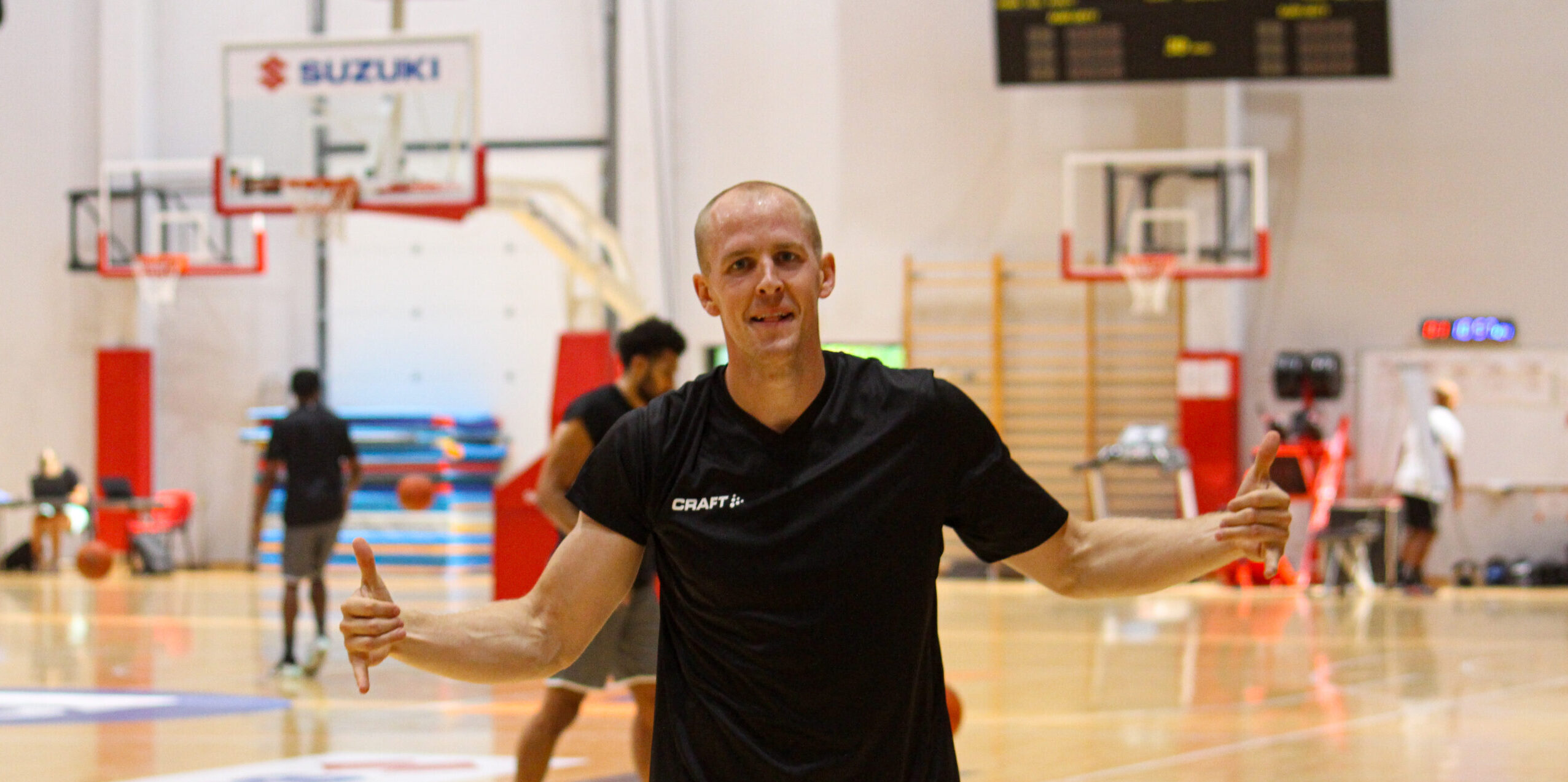 Mann in schwarzem T-Shirt zeigt Daumen nach oben in einer Basketballhalle mit Körben im Hintergrund