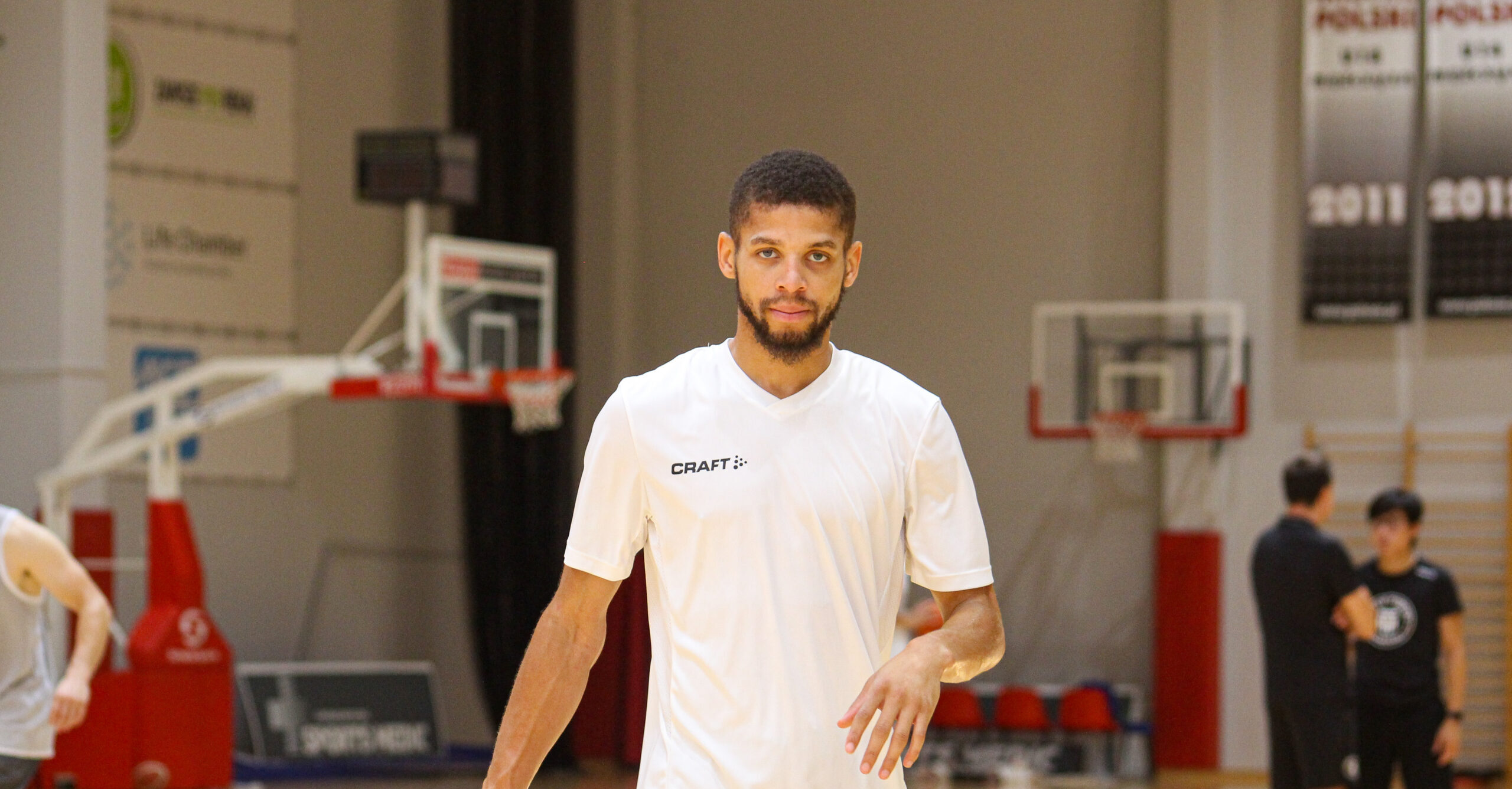 Junger Mann in weißem T-Shirt steht in einer Basketballhalle mit Körben im Hintergrund