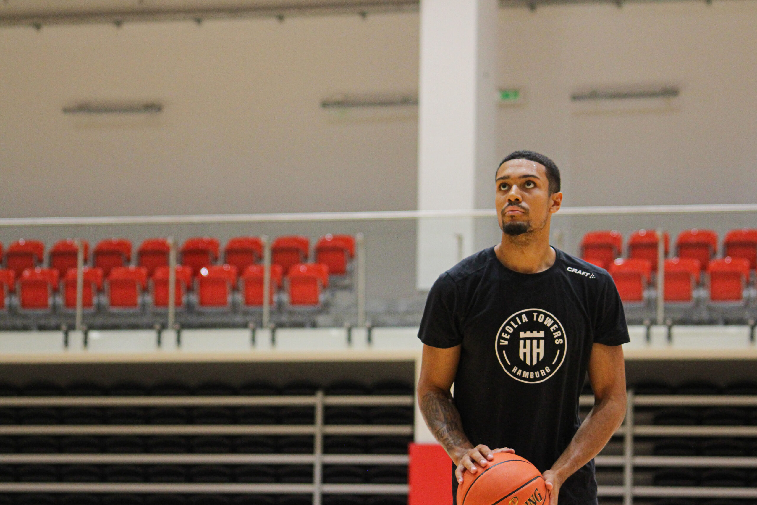 Basketballspieler in schwarzem T-Shirt mit Ball in der Hand in einer leeren Sporthalle mit roten Sitzen