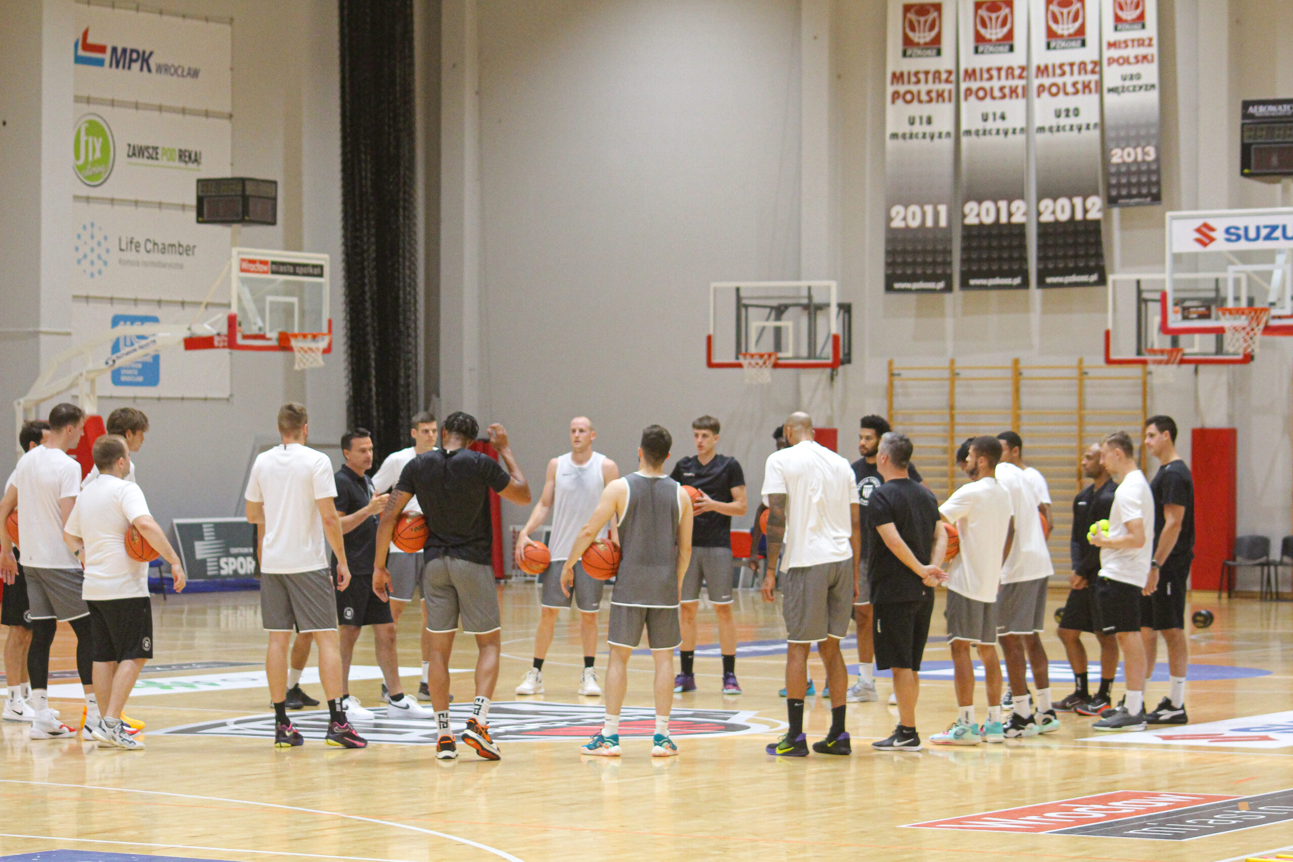 Basketballspieler und Trainer stehen im Kreis auf einem Hallenboden, einige halten Basketbälle, Trainingsumgebung mit Körben und Bannern im Hintergrund