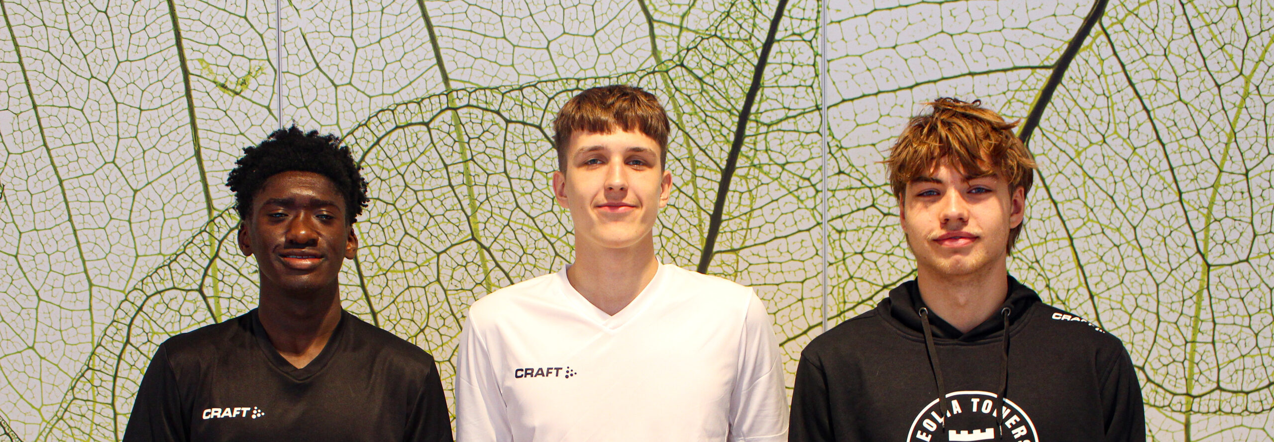 Drei junge Männer stehen nebeneinander vor einem Hintergrund mit einem großen, grünen Blattmuster. Sie tragen sportliche Kleidung von Craft.