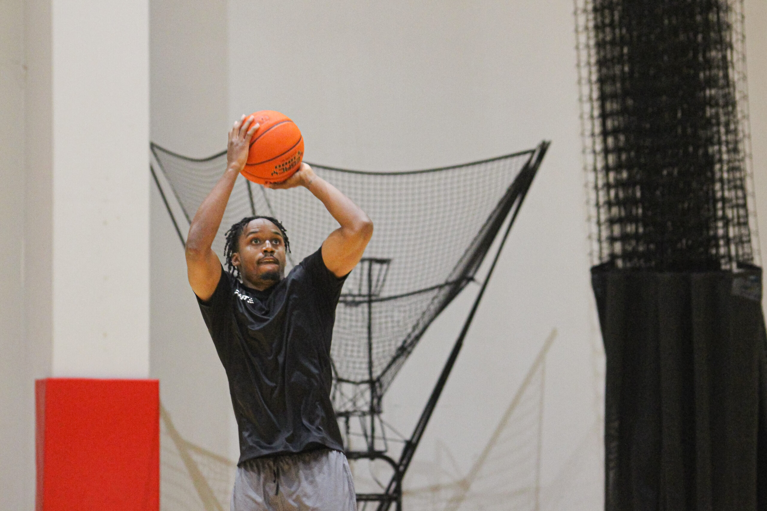 Basketballspieler in schwarzem Shirt hebt den Ball zum Wurf in einer Halle mit Basketballnetz im Hintergrund