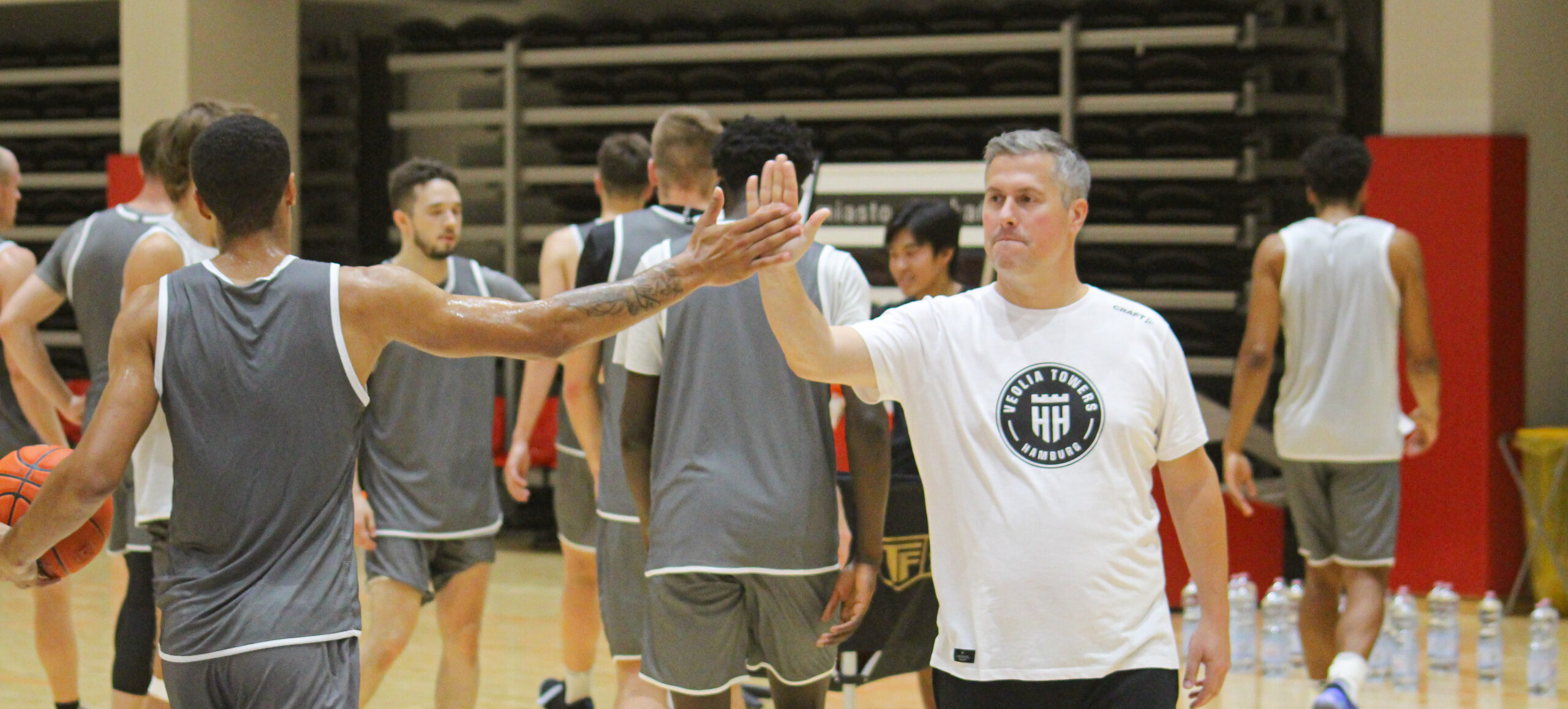Basketballspieler und Trainer geben sich in der Halle einen High-Five, Spieler tragen graue Trikots, Trainer weißes T-Shirt mit Logo