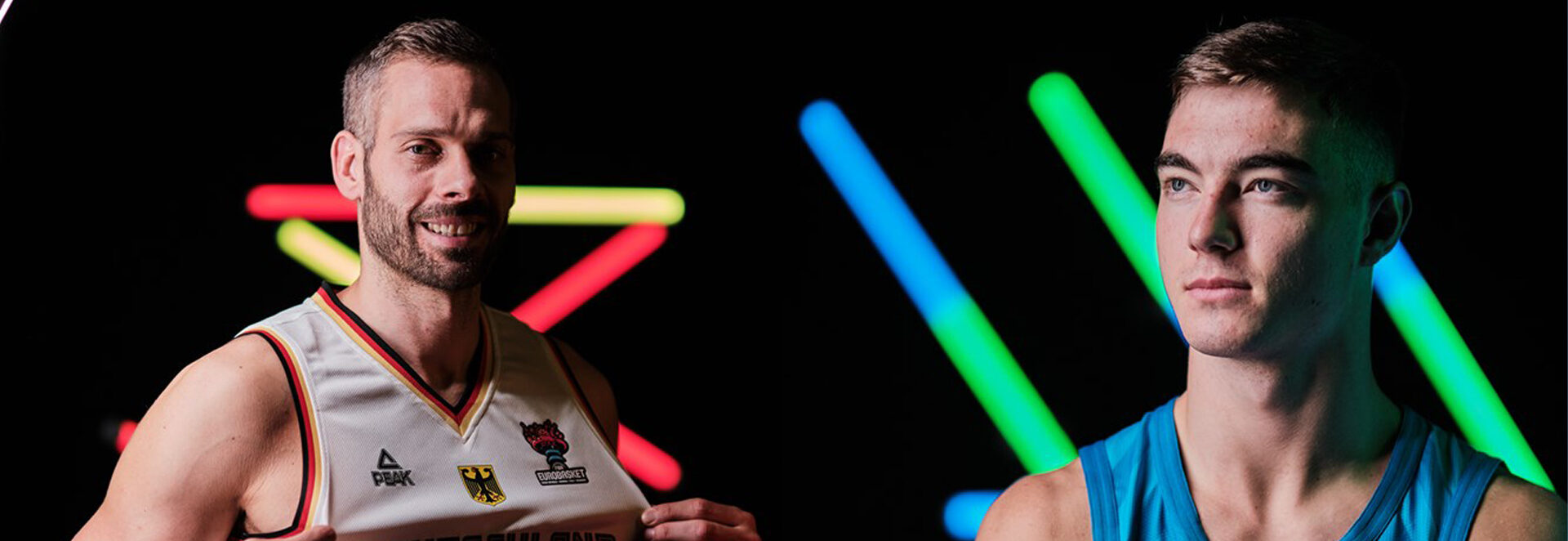 Zwei männliche Basketballspieler vor schwarzem Hintergrund mit bunten Neonlichtern, einer trägt weißes Trikot, der andere blaues Trikot