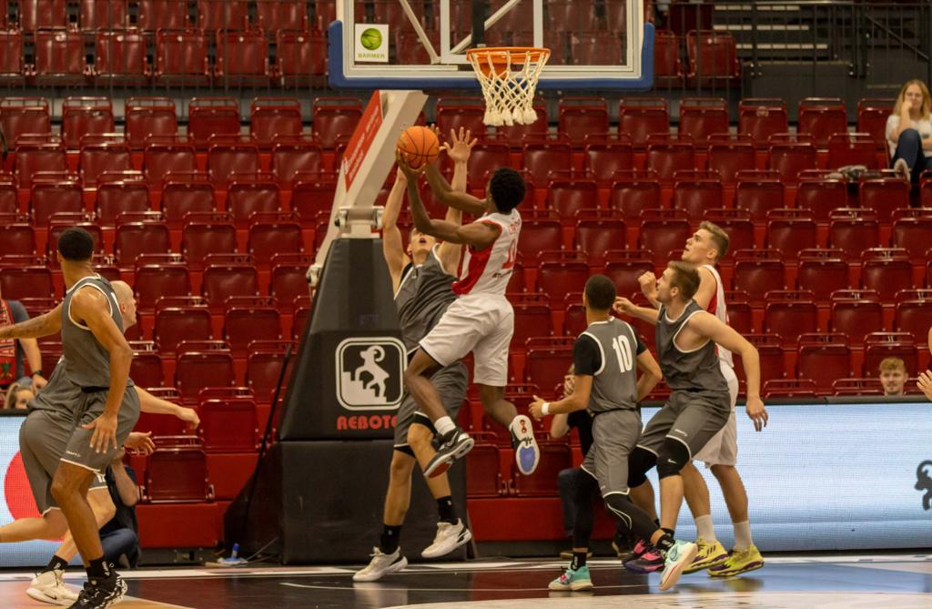 Basketballspieler in weißem Trikot versucht Korb zu erzielen, während Spieler in grauen Trikots verteidigen, leere Tribünen im Hintergrund