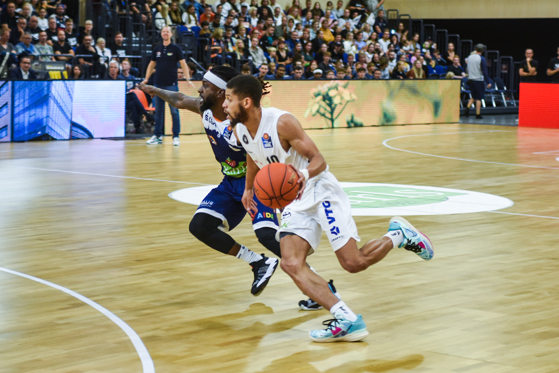 Zwei Basketballspieler in Aktion auf dem Spielfeld, einer in weiß dribbelt den Ball, der andere in dunkelblau verteidigt, Zuschauer im Hintergrund