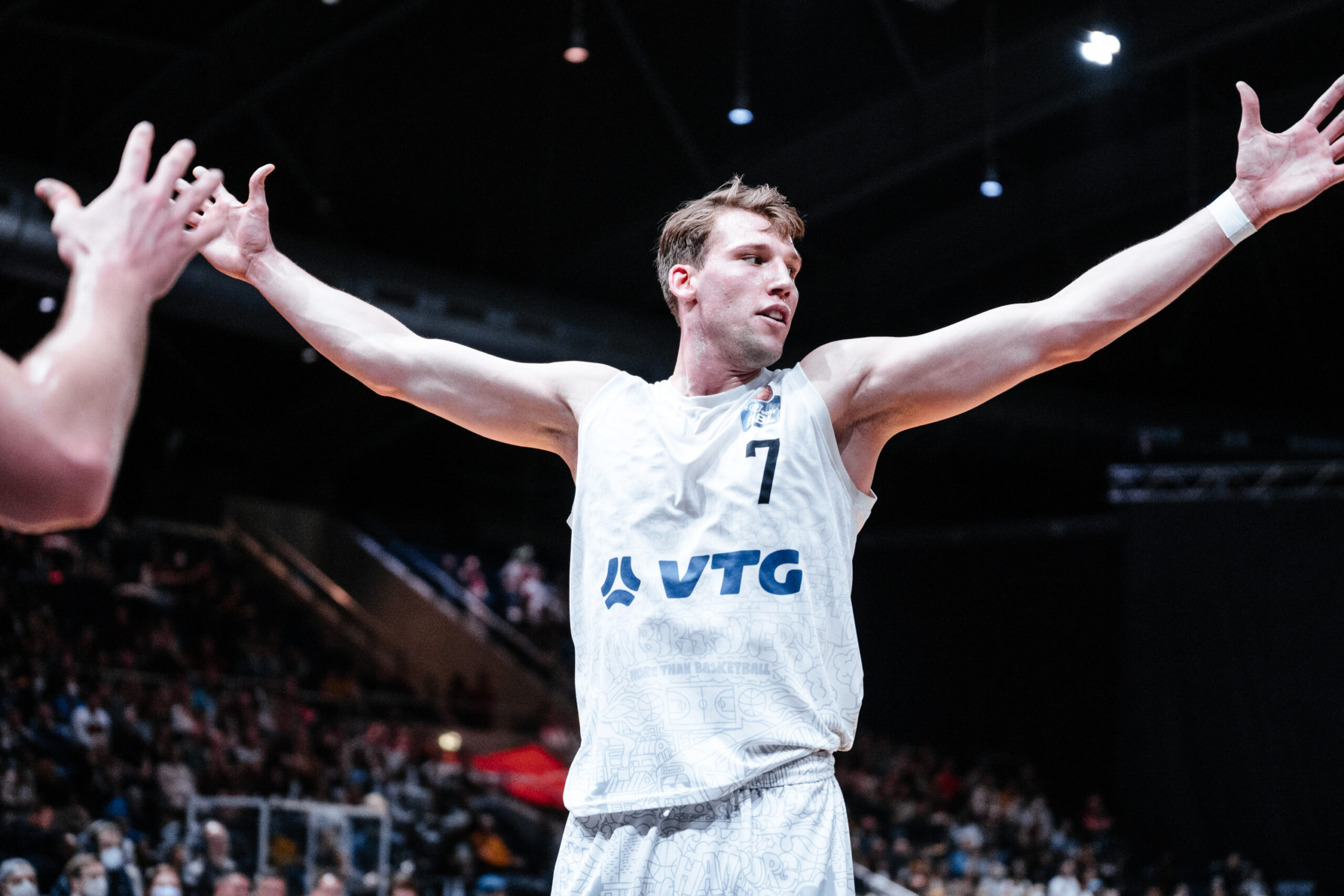 Basketballspieler mit weißem Trikot Nummer 7 und 'VTG'-Logo breitet die Arme aus, im Hintergrund Zuschauer in der Halle