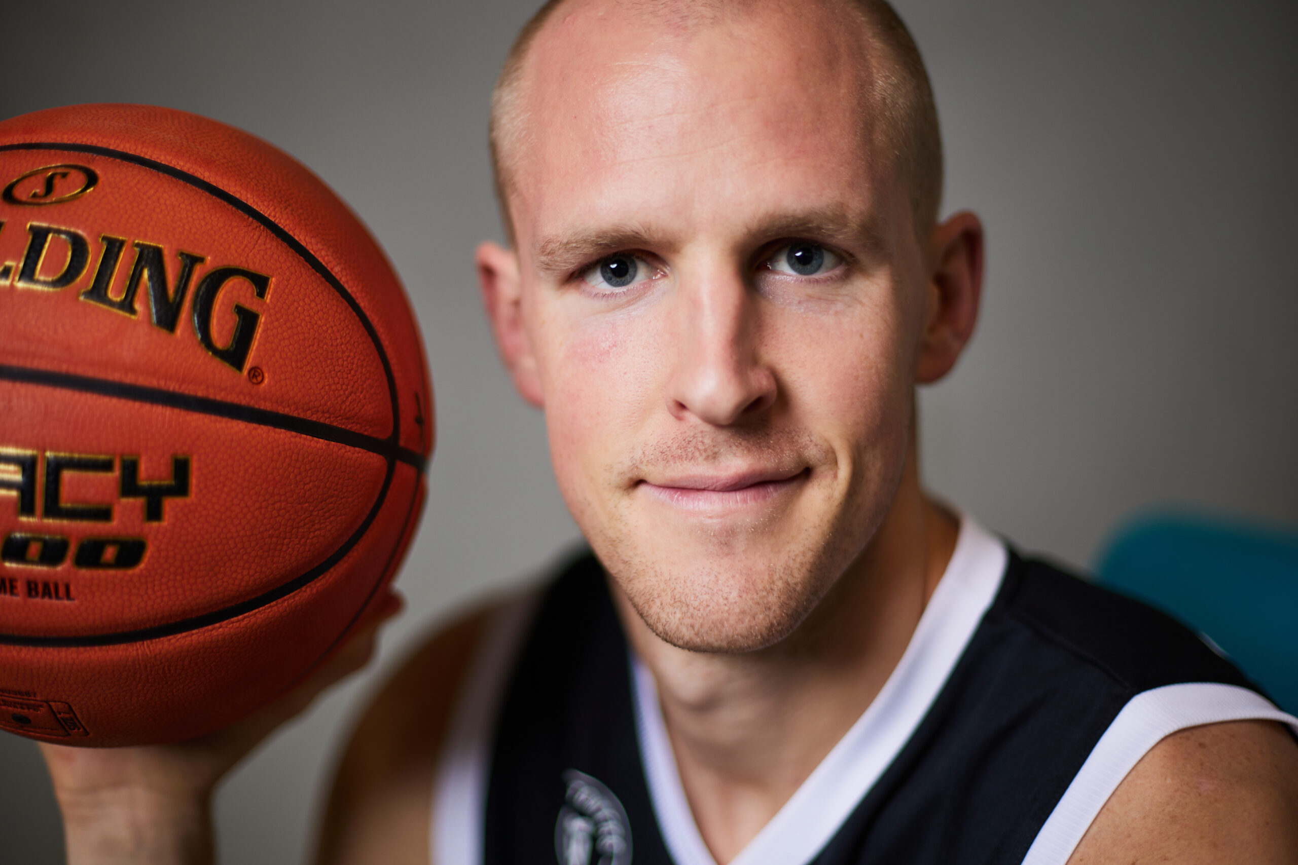 Nahaufnahme eines Mannes mit kurzem blondem Haar, der einen orangefarbenen Spalding-Basketball hält und in schwarzem Basketballtrikot lächelt