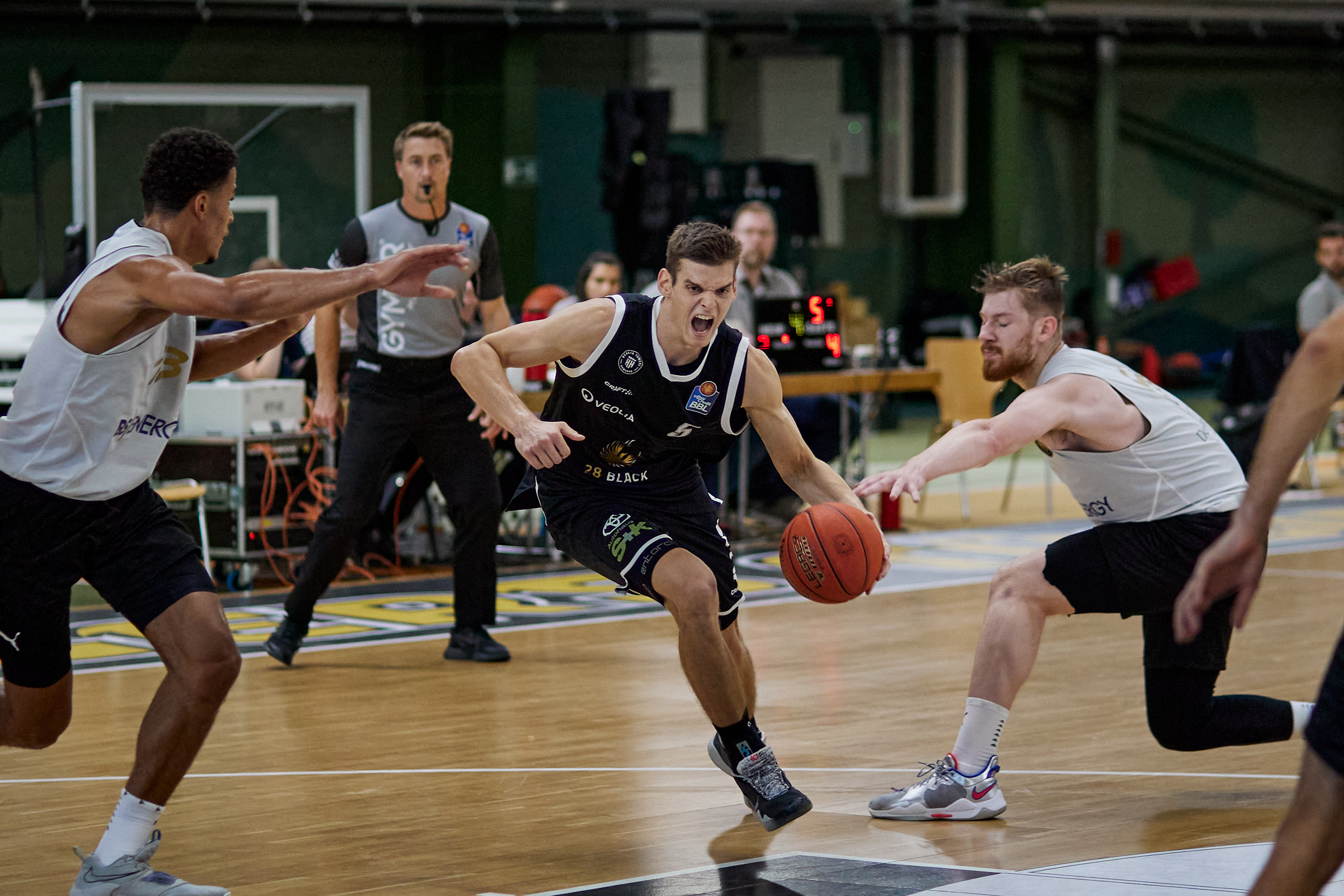 Basketballspieler in schwarzem Trikot dribbelt energisch, während zwei Spieler in weißen Trikots versuchen, ihn zu blockieren