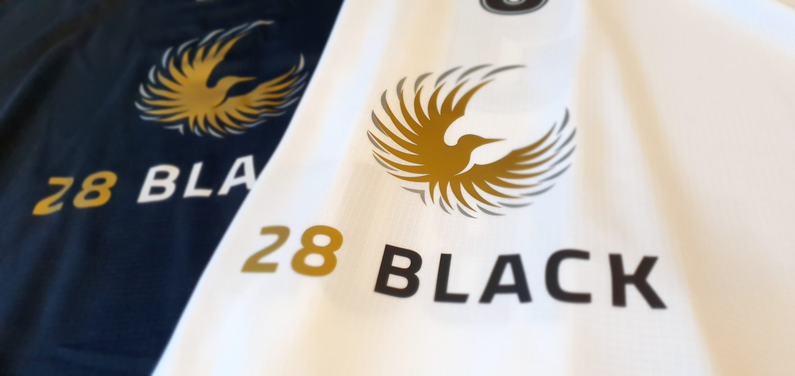 Zwei Textilien mit dem goldenen Logo eines stilisierten Vogels und dem Schriftzug '28 BLACK', eines in Schwarz, das andere in Weiß